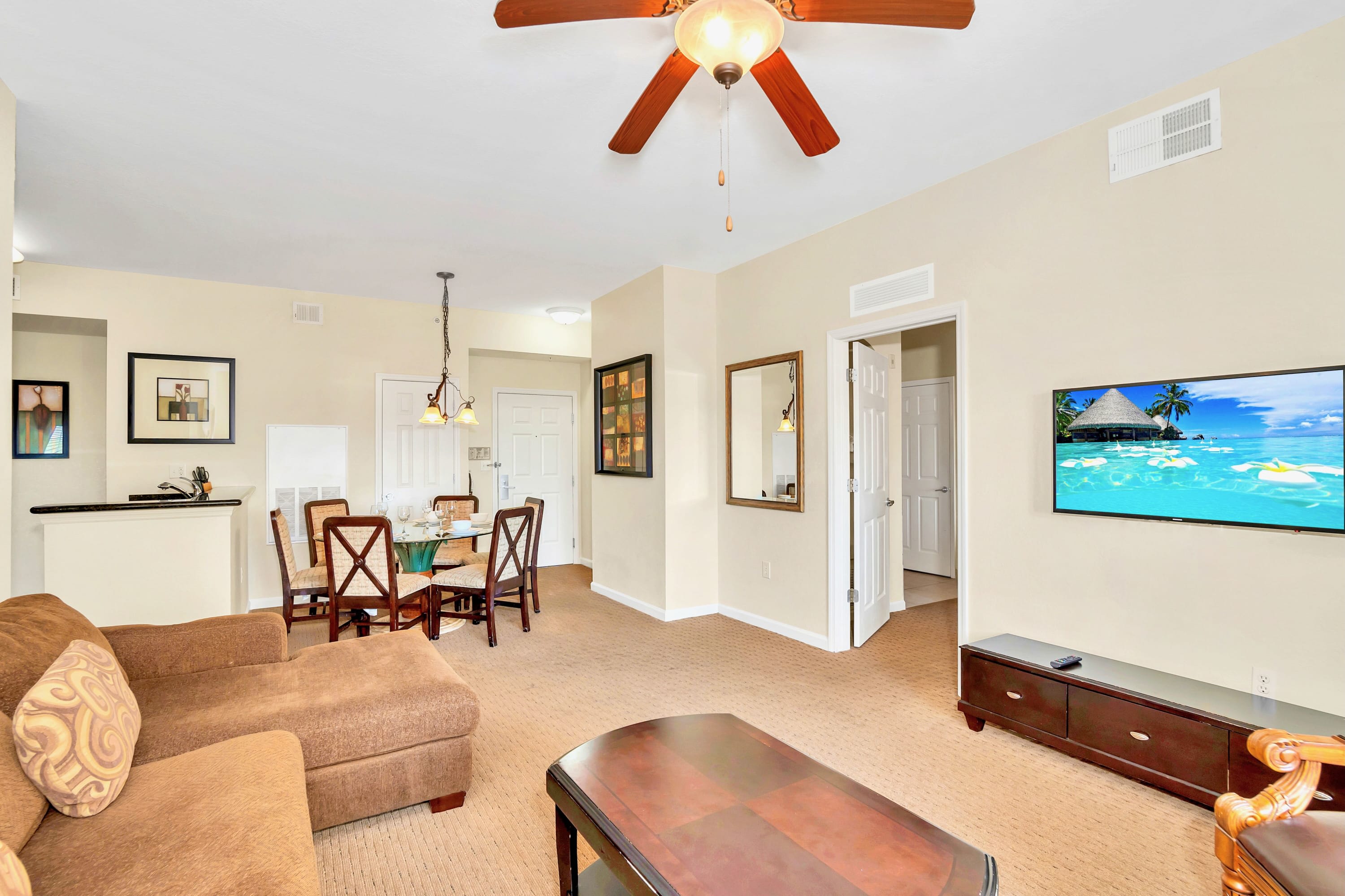 An open-concept living area features a brown sectional sofa, a dark wood coffee table, and a wall-mounted TV. A dining area with a glass-top table and chairs is visible in the background, along with a hallway.