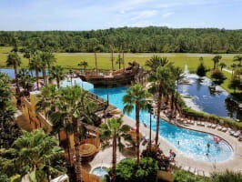 An aerial shot reveals a large, irregularly shaped swimming pool complex featuring a prominent pirate ship structure with water slides. Lush tropical foliage and palm trees surround the pool, with resort buildings visible in the background.
