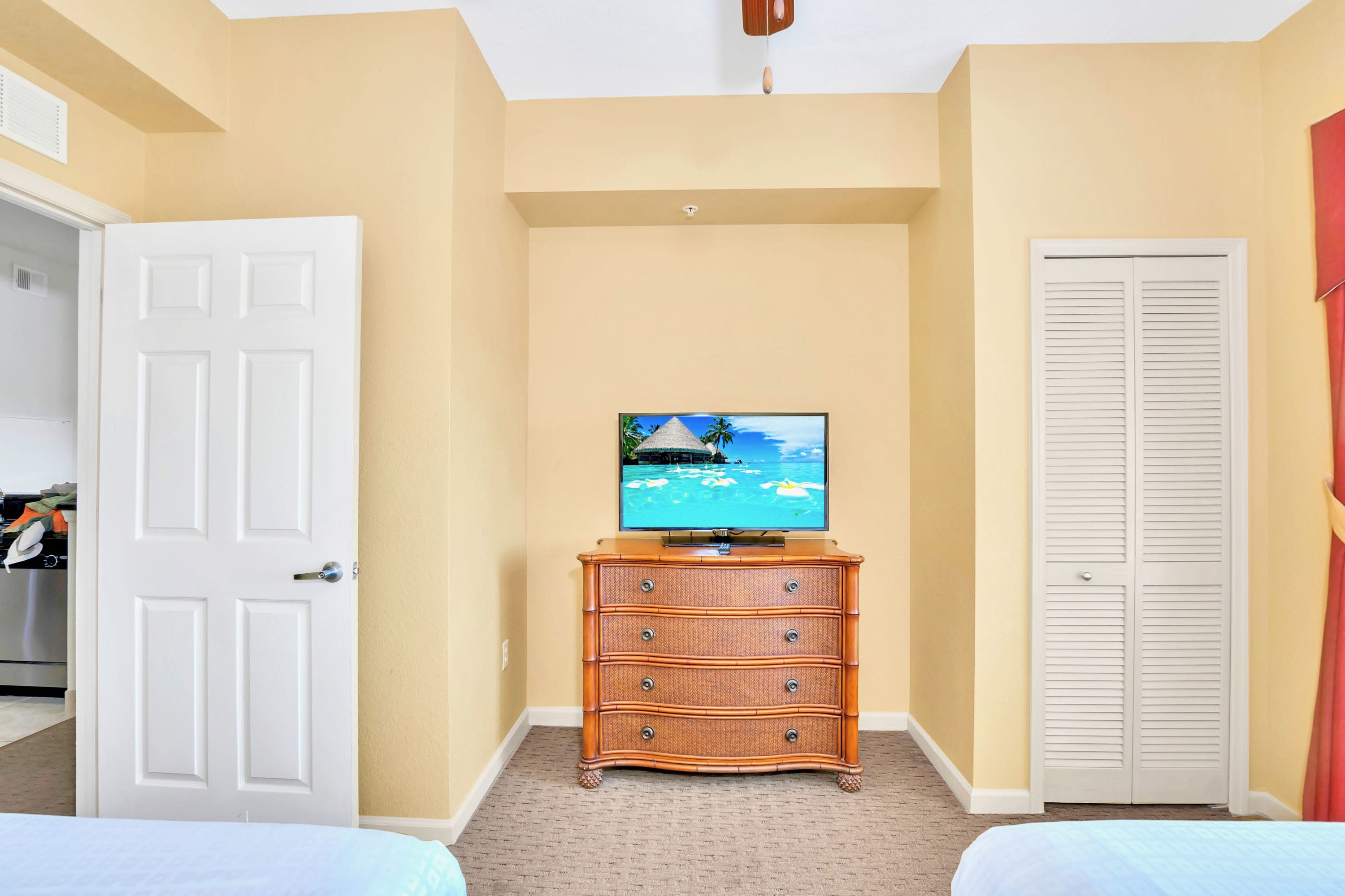 View of the second bedroom showing a wicker dresser with a television, a closed bi-fold closet, and a doorway leading to another room, with two beds partially visible.