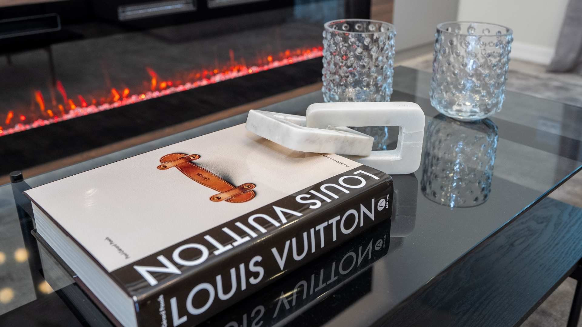 Close-up of a black glass coffee table in the living room, adorned with a "Louis Vuitton" book and two clear glass candle holders. The table sits on a grey area rug, with the modern electric fireplace glowing in the background.