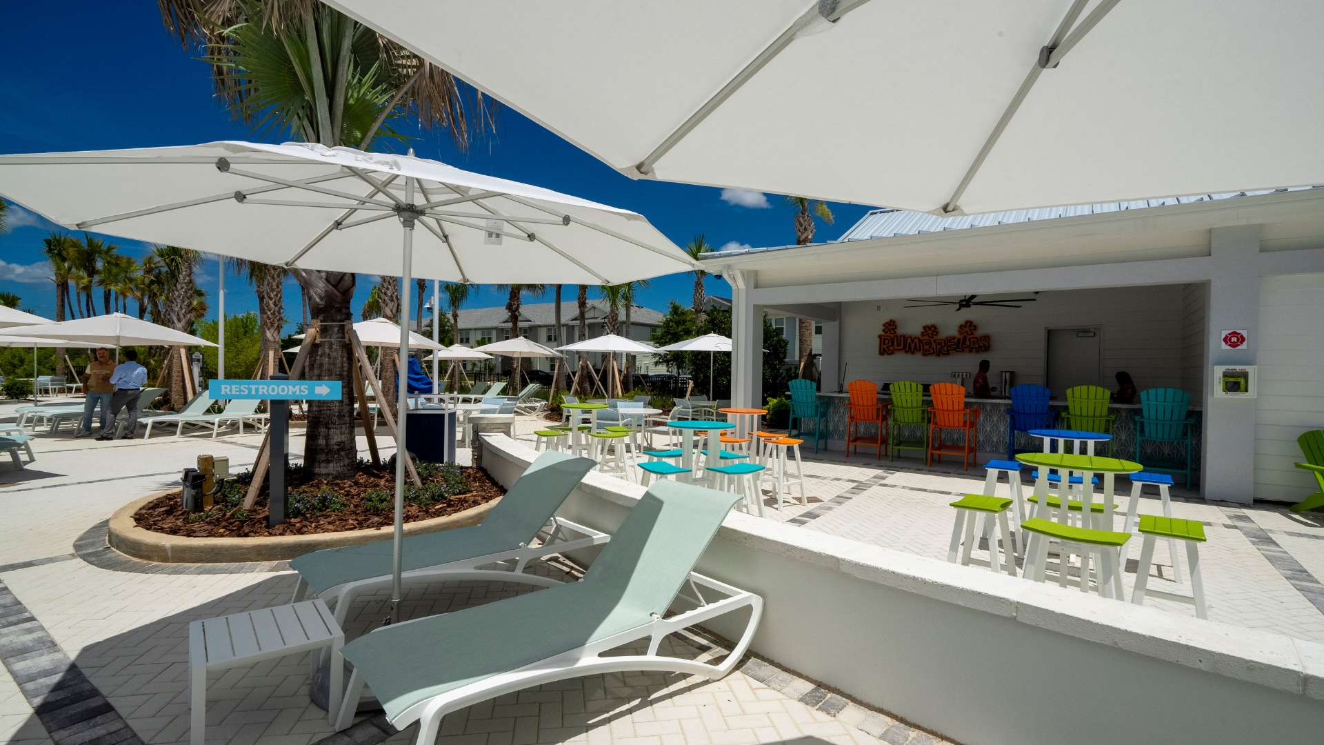 A vibrant outdoor bar area with a white counter, colorful bar stools, and tables, shaded by large white umbrellas, with a 'RESTROOMS' sign indicating nearby facilities.
