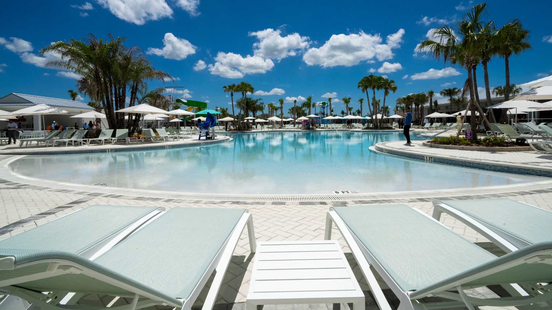 A wide-angle view of the large, light-blue swimming pool with a shallow entry, surrounded by a spacious white paved deck filled with numerous lounge chairs and white umbrellas, under a clear blue sky.