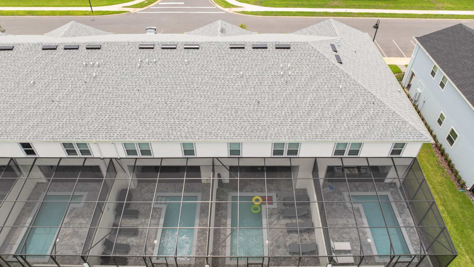 An aerial shot directly above a row of townhouses reveals multiple private screened-in pools in their backyards, each with lounge chairs and some with floating toys. The roofs are light gray shingles, and the surrounding area is neatly landscaped.