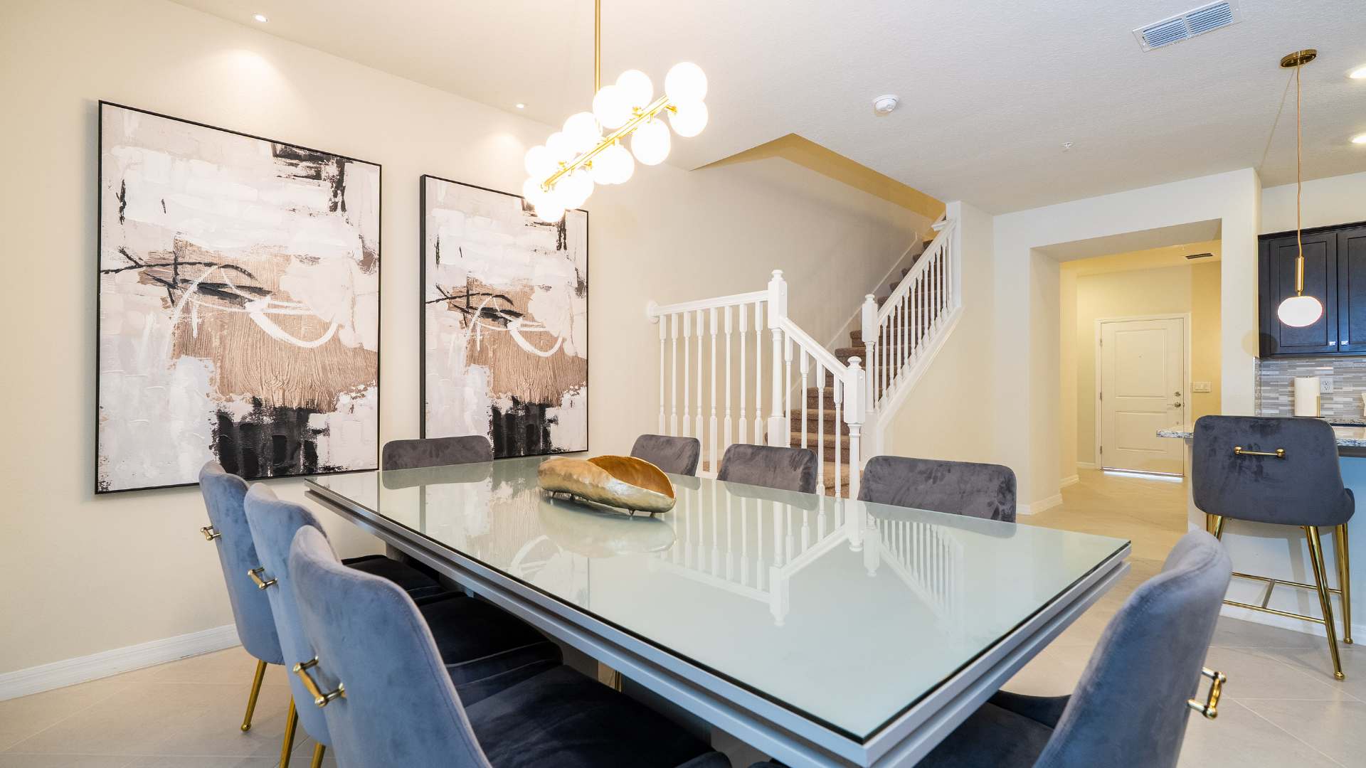 Modern dining room featuring a long glass-top table with dark velvet chairs, illuminated by a linear gold chandelier, and abstract art on the wall. A white staircase with a gold accent is visible in the background.