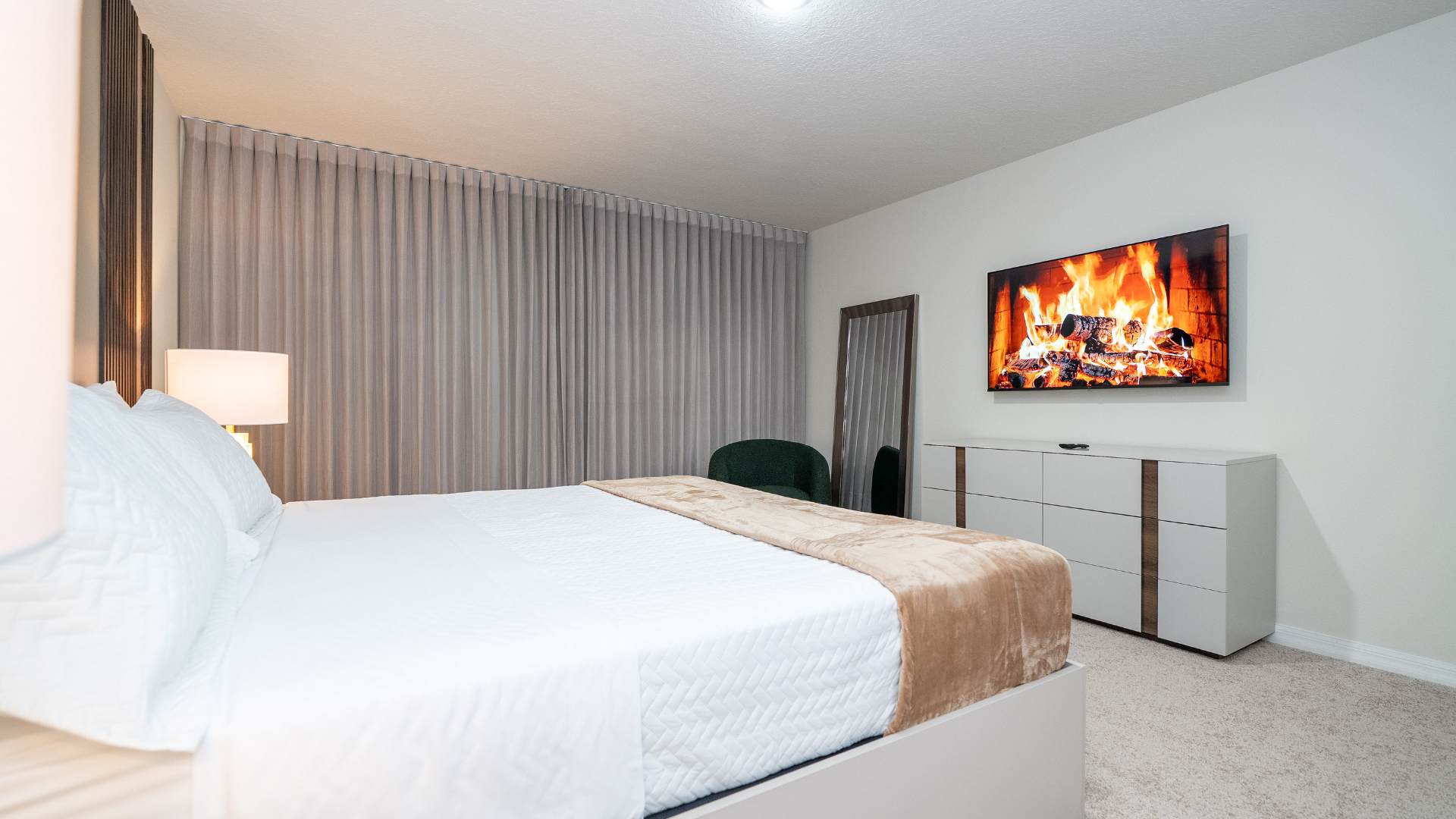 The bedroom features a wall-mounted flat-screen TV displaying a fireplace scene, positioned above a white dresser with wooden accents. A full-length mirror leans against the wall, and a dark green textured accent chair sits nearby.