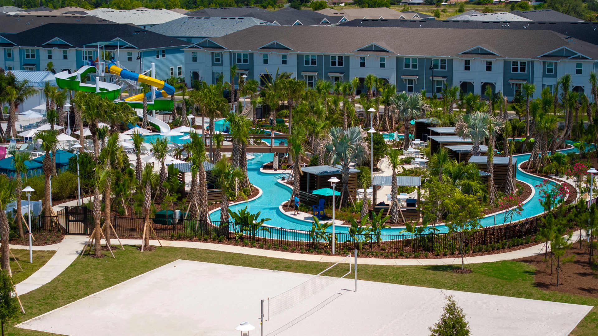 An aerial view showcasing the entire resort water park, including a winding lazy river, large swimming pools, colorful water slides, and numerous palm trees, with a sand volleyball court in the foreground and residential buildings in the background.