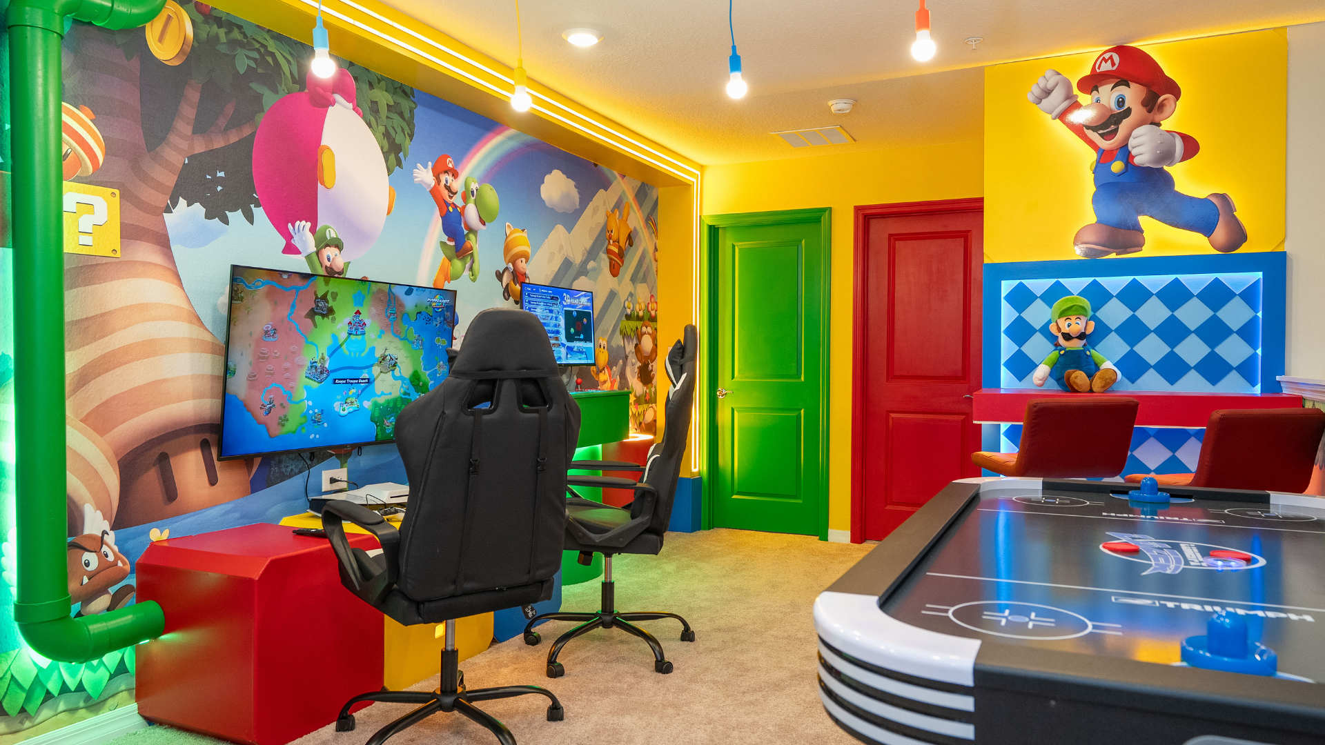 A brightly lit game room showcases a large TV displaying a video game, two gaming chairs, and an air hockey table. The walls are adorned with extensive Mario-themed murals, and colorful doors lead to other areas, creating an immersive play space.