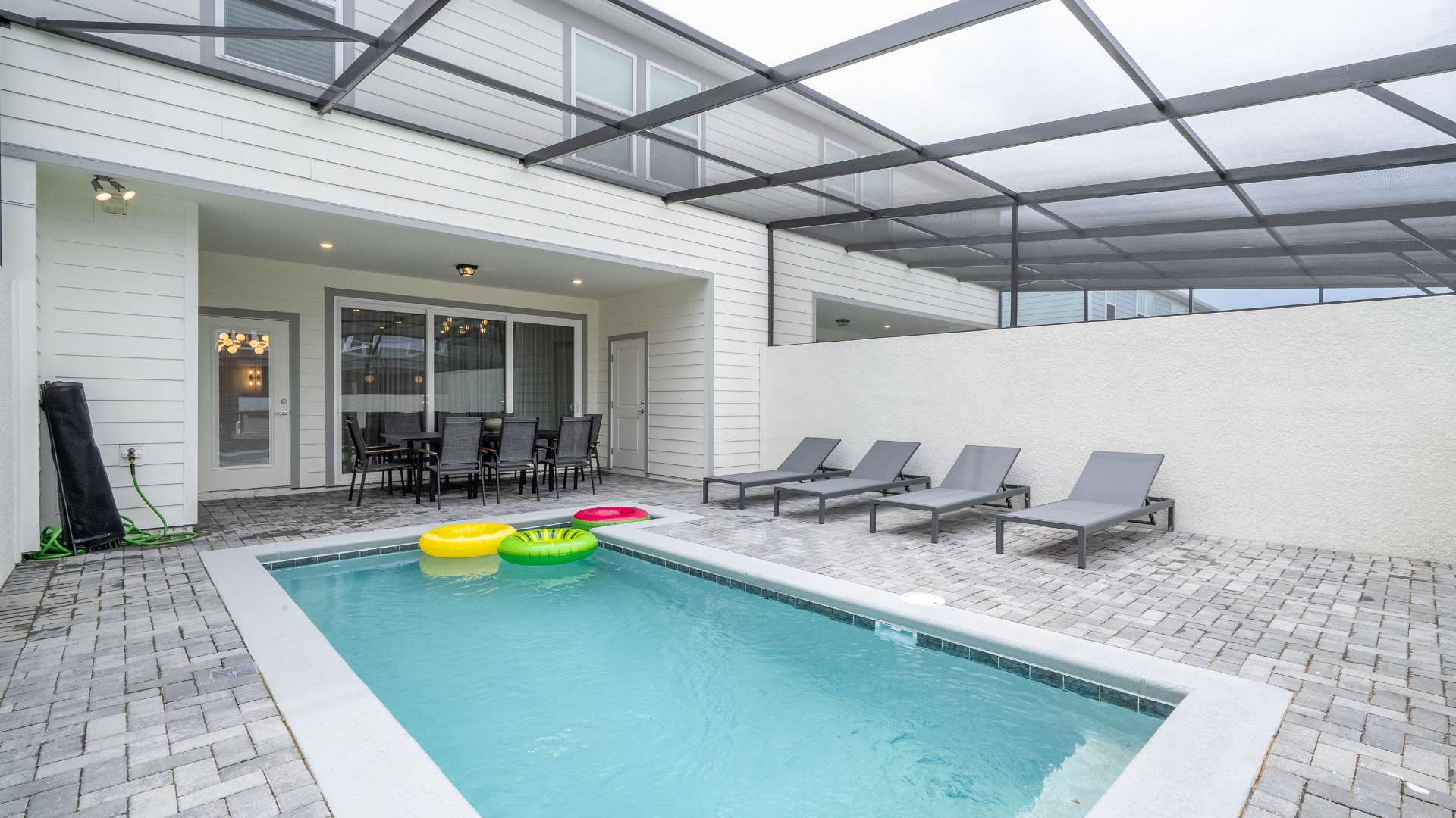 An outdoor private swimming pool is surrounded by a gray brick patio and enclosed by a screen cage, with four gray lounge chairs positioned along one side. Colorful inflatable rings float in the clear blue water, and the house exterior is visible.