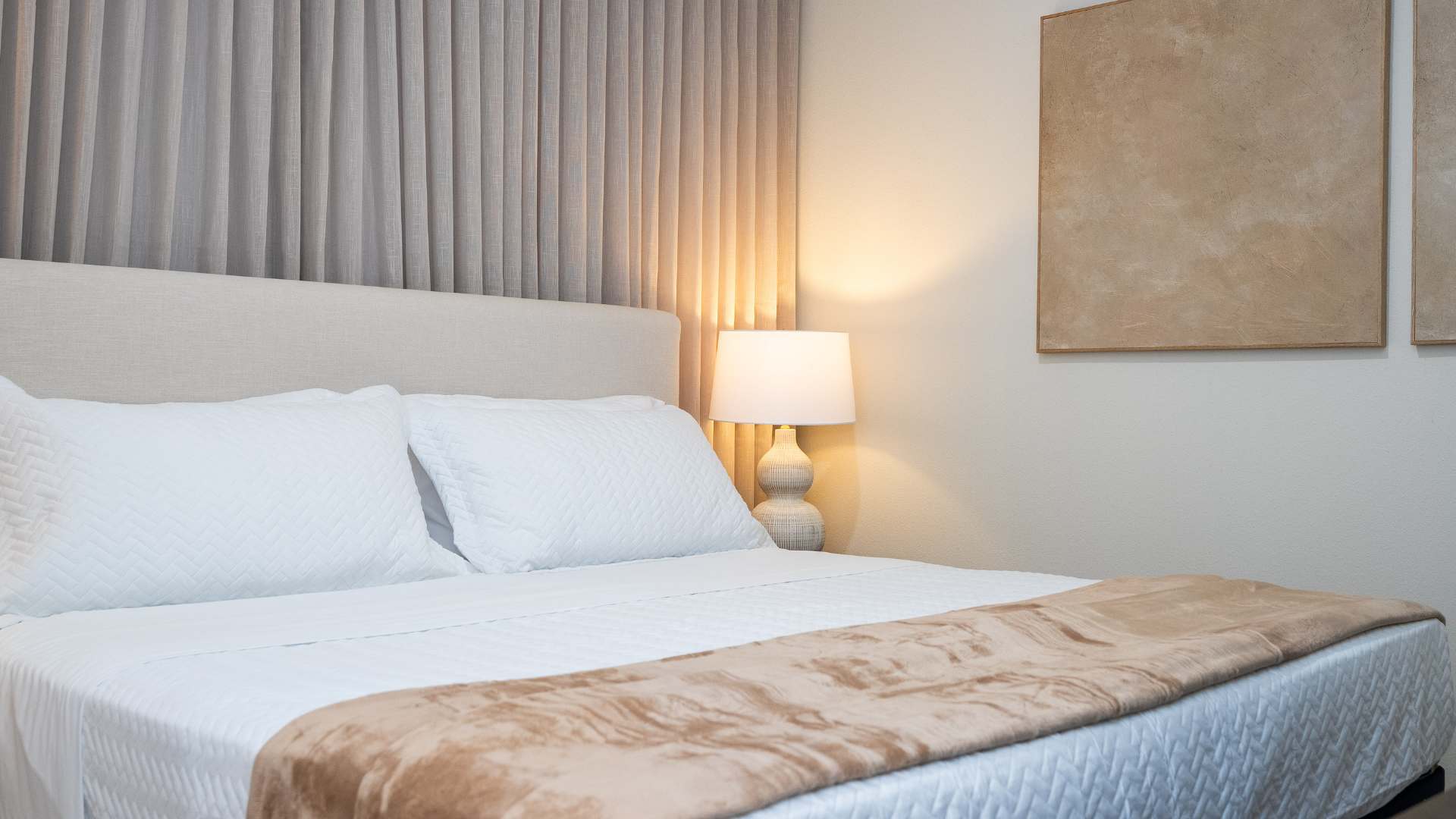 Close-up of a bed with white quilted bedding and a tan throw, featuring a light beige upholstered headboard and a nightstand with a textured ceramic lamp.