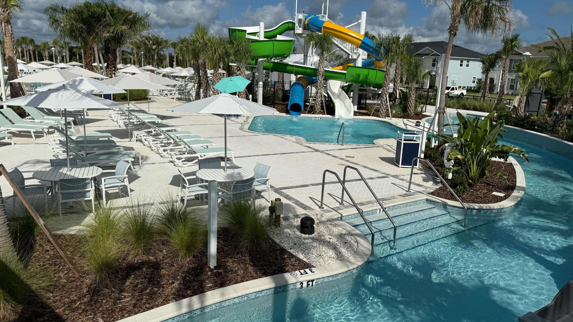 Bright and inviting communal pool area showcasing a large water slide complex, a curved lazy river, and a wide paved sun deck with numerous lounge chairs and white shade umbrellas.