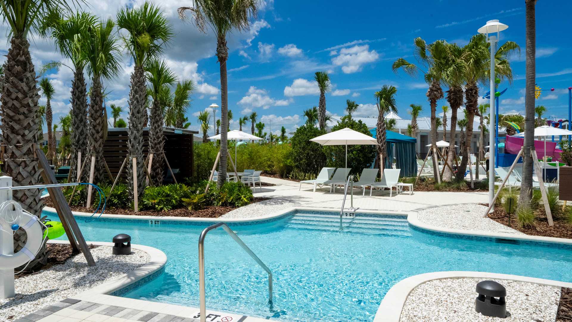 A serene section of the lazy river with steps and a metal handrail for entry, bordered by white gravel, lush greenery, and palm trees, with lounge chairs and white umbrellas in the background.