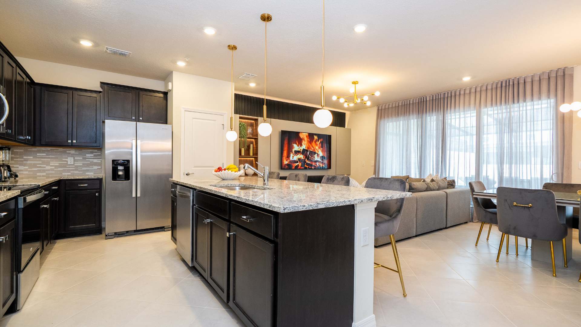 Spacious open-concept kitchen and living area with a large granite island, dark cabinetry, and stainless steel appliances. Gold pendant lights illuminate the island, with a view of the living room's large TV and dining area beyond.