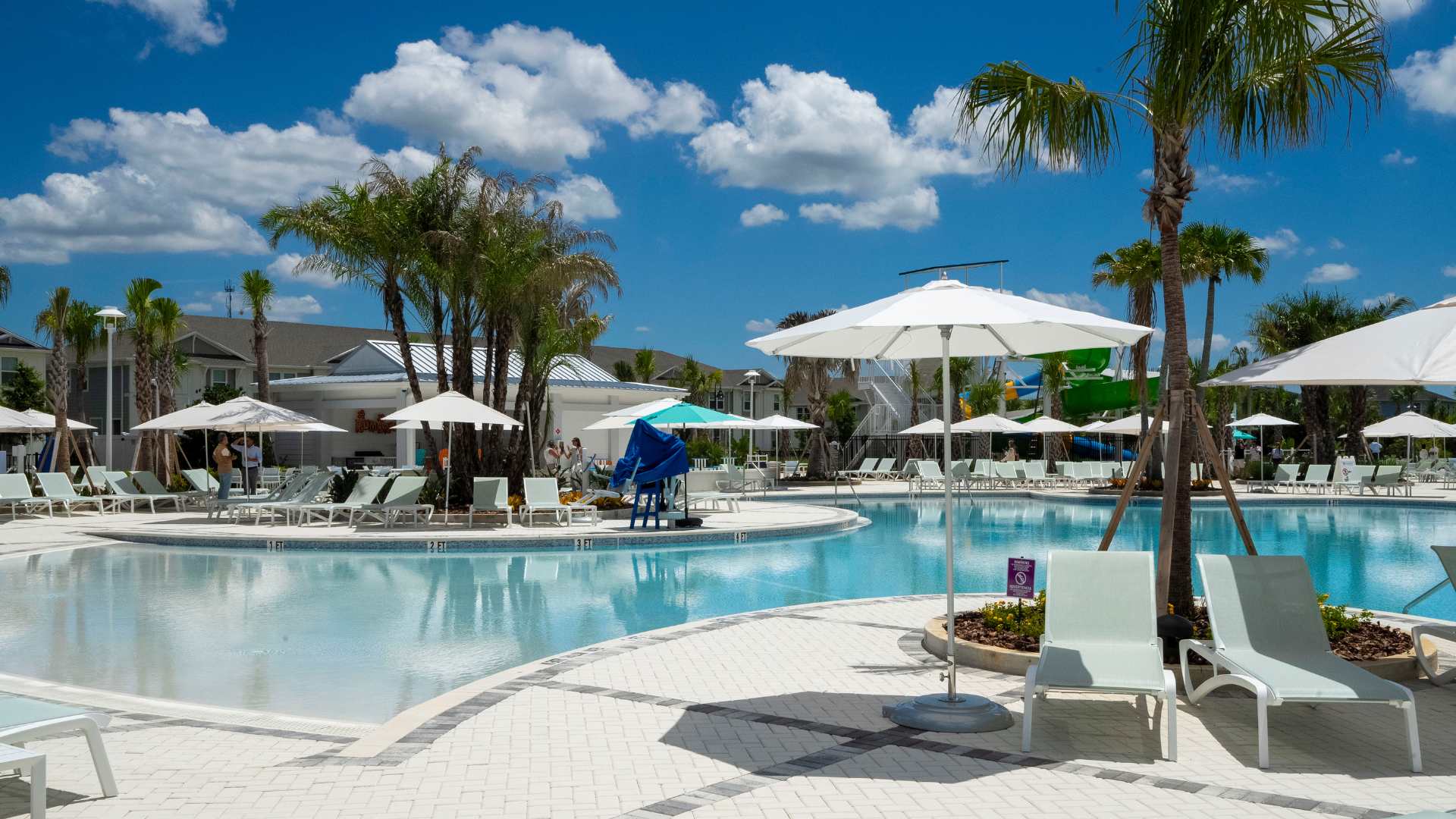 A expansive main swimming pool with a gradual zero-entry slope, surrounded by a wide paved deck furnished with numerous white lounge chairs and large white umbrellas, under a bright blue sky.