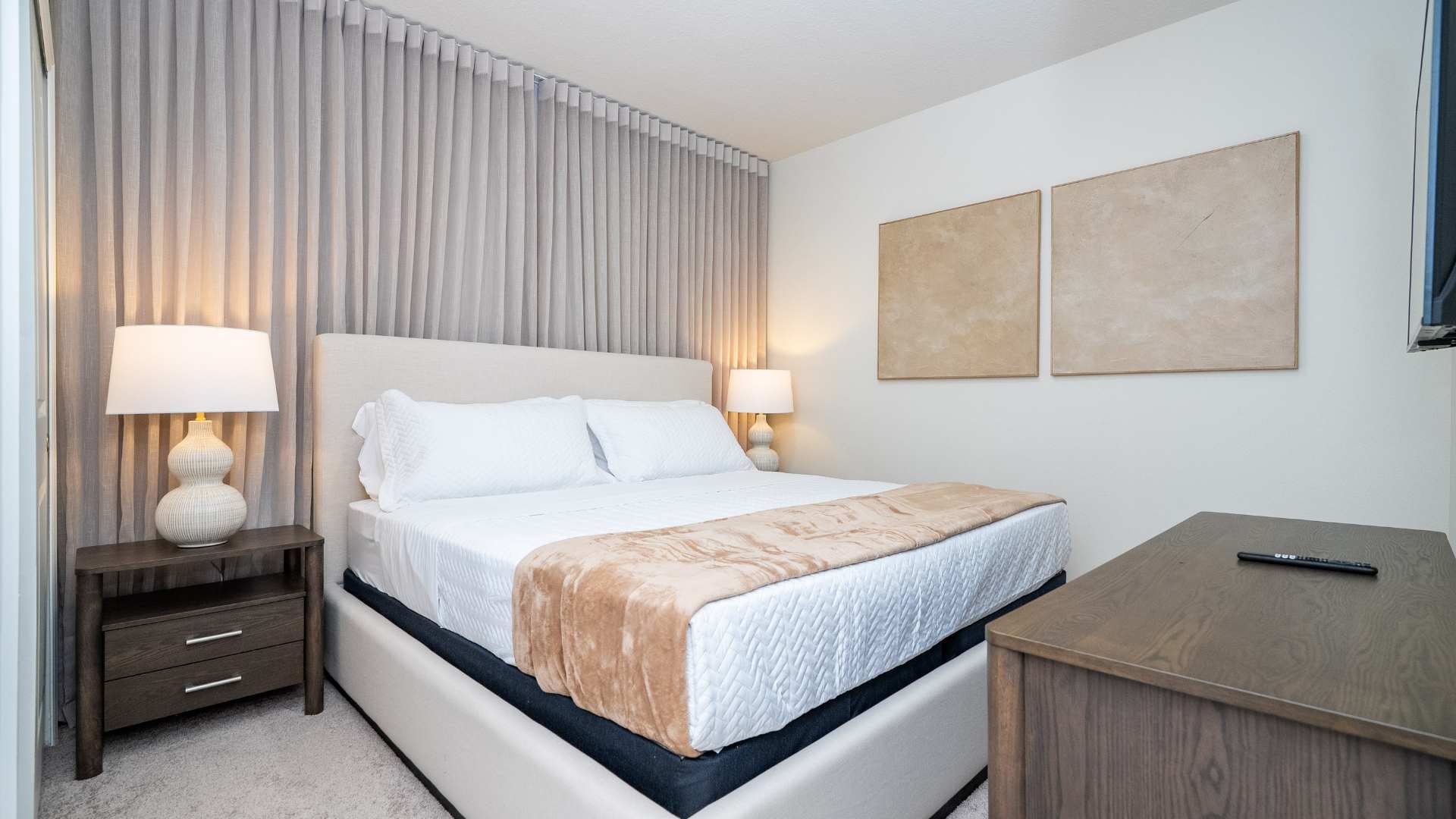 Wide shot of a bedroom showcasing a light beige upholstered bed with white and tan bedding, flanked by dark wood nightstands and lamps, with two abstract art pieces on the adjacent wall.