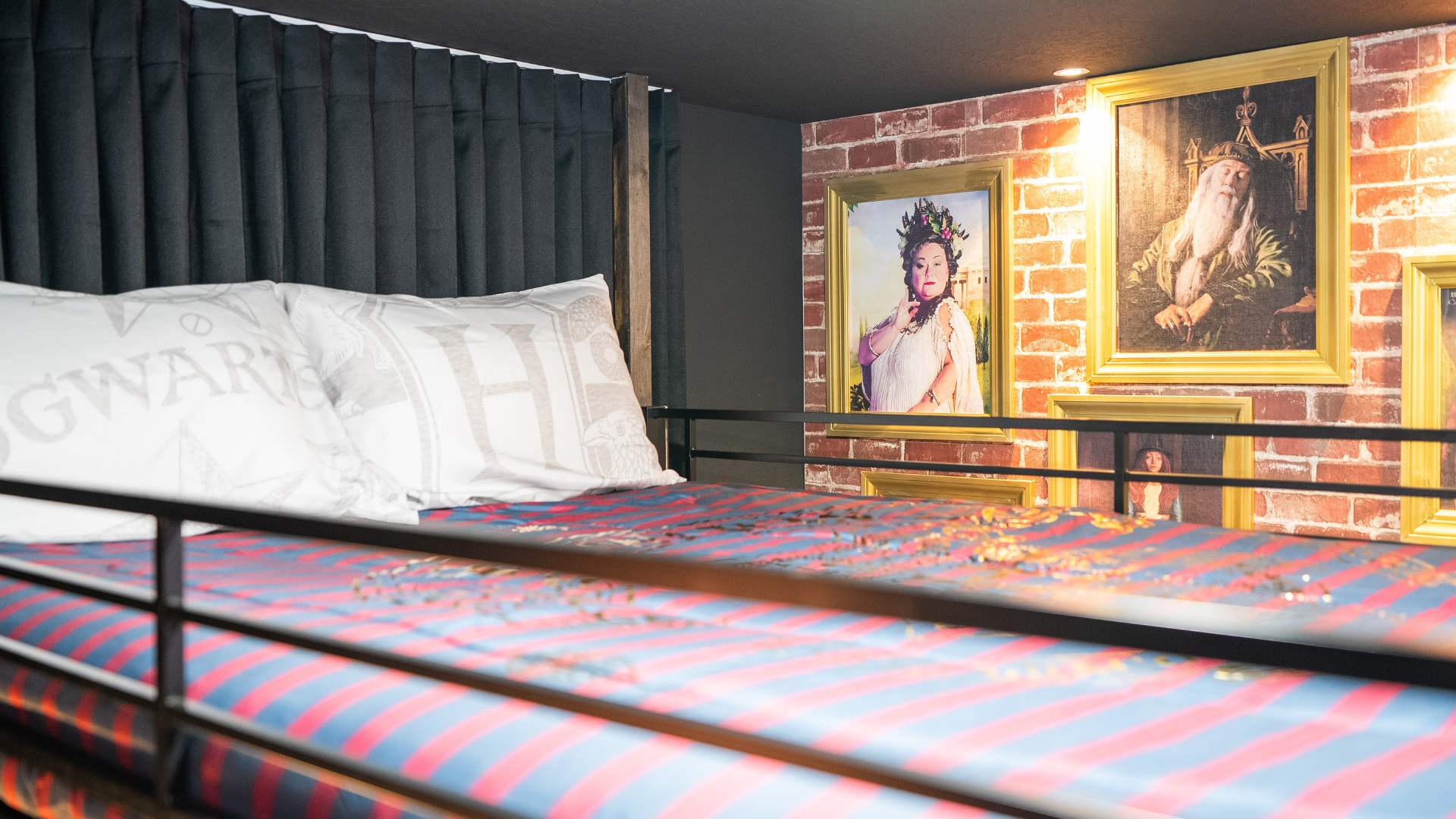 Upper bunk of the Harry Potter themed bedroom, featuring "Hogwarts" pillows and a wall adorned with framed portraits, including Dumbledore, against a brick background.