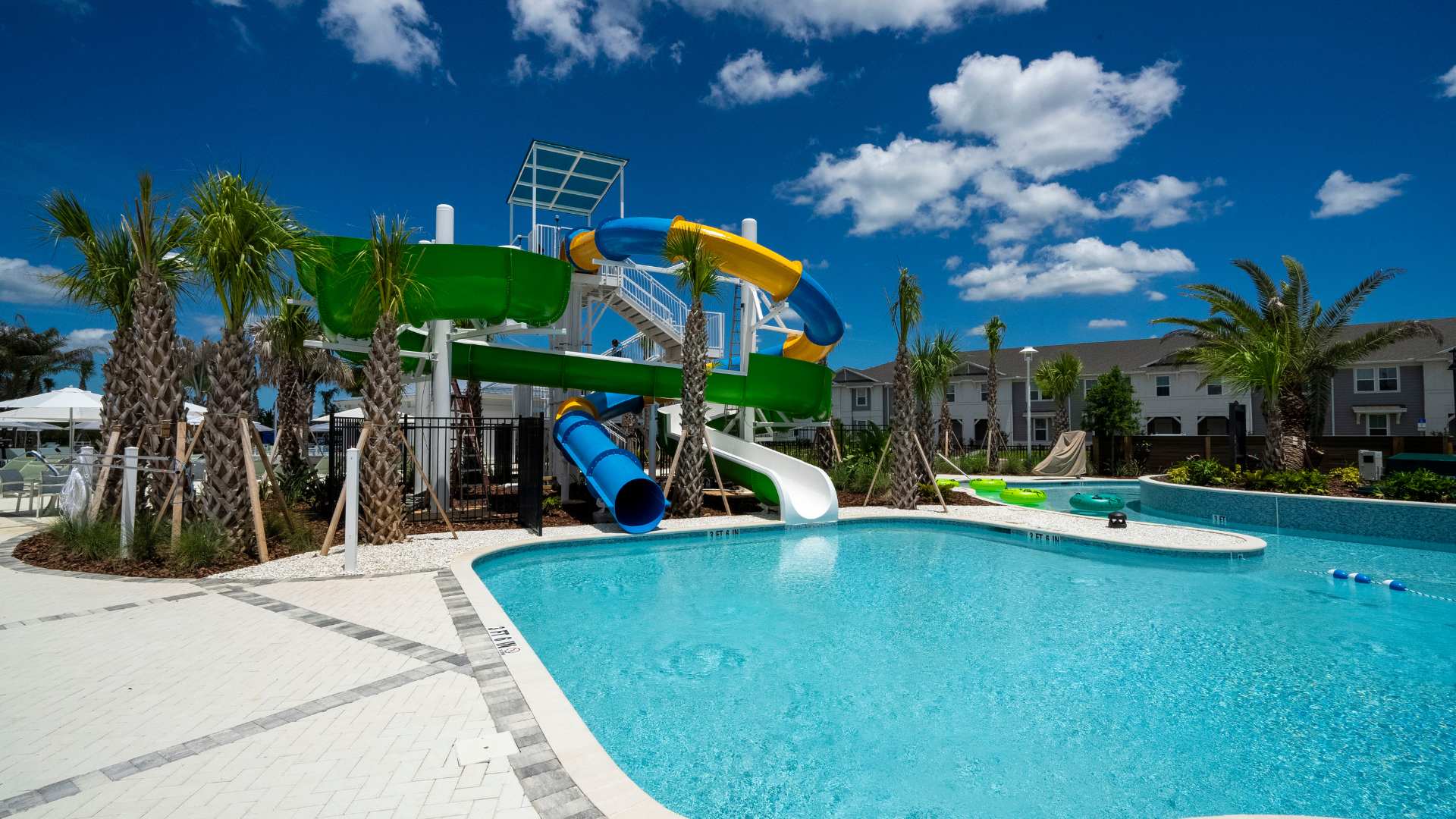 A large, inviting swimming pool with a vibrant multi-story water slide featuring blue, yellow, green, and white slides, surrounded by palm trees and resort-style buildings under a bright blue sky.