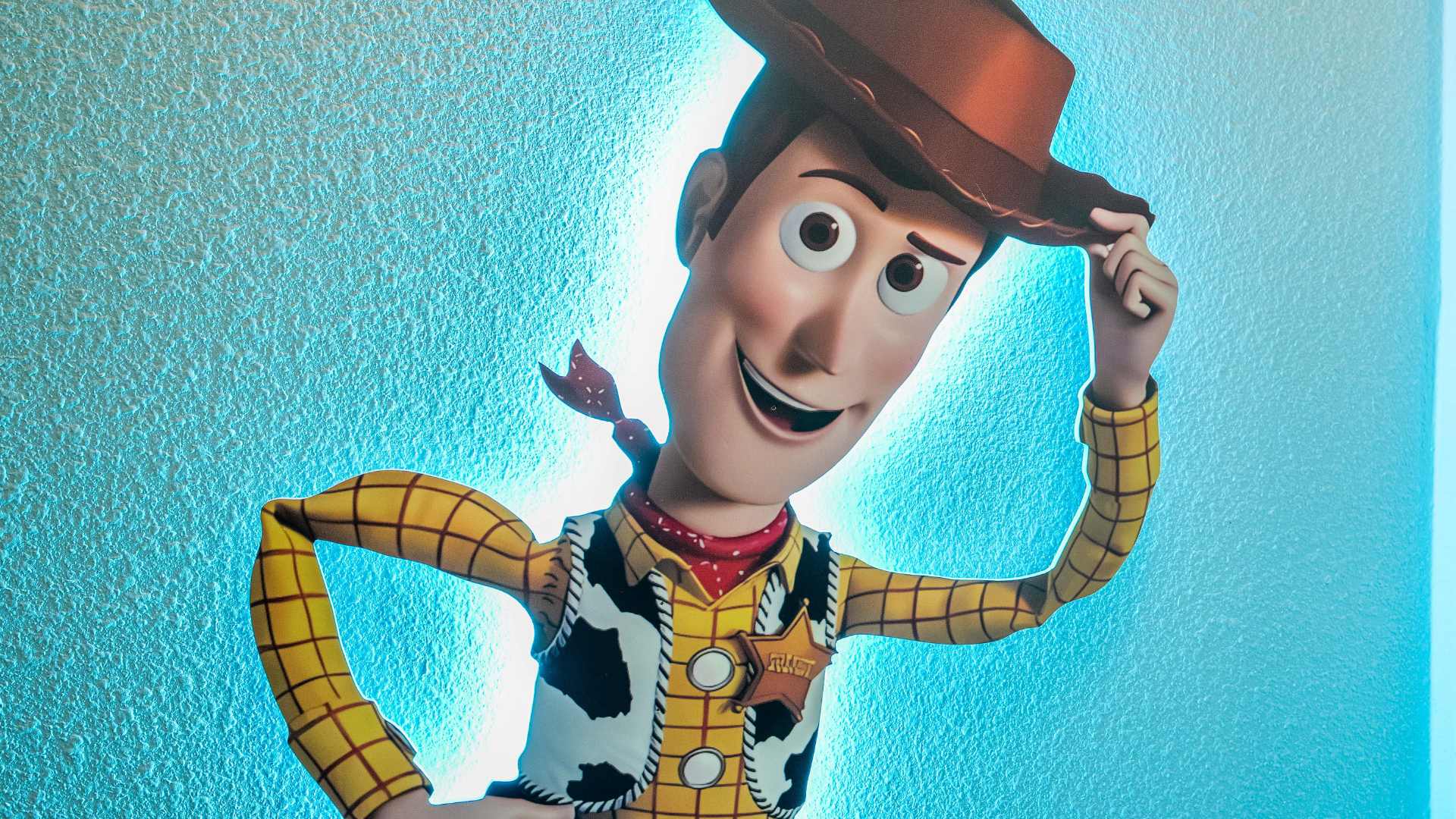 A large, vibrant graphic of Woody from Toy Story, smiling and tipping his hat, set against a textured blue wall with bright backlighting.