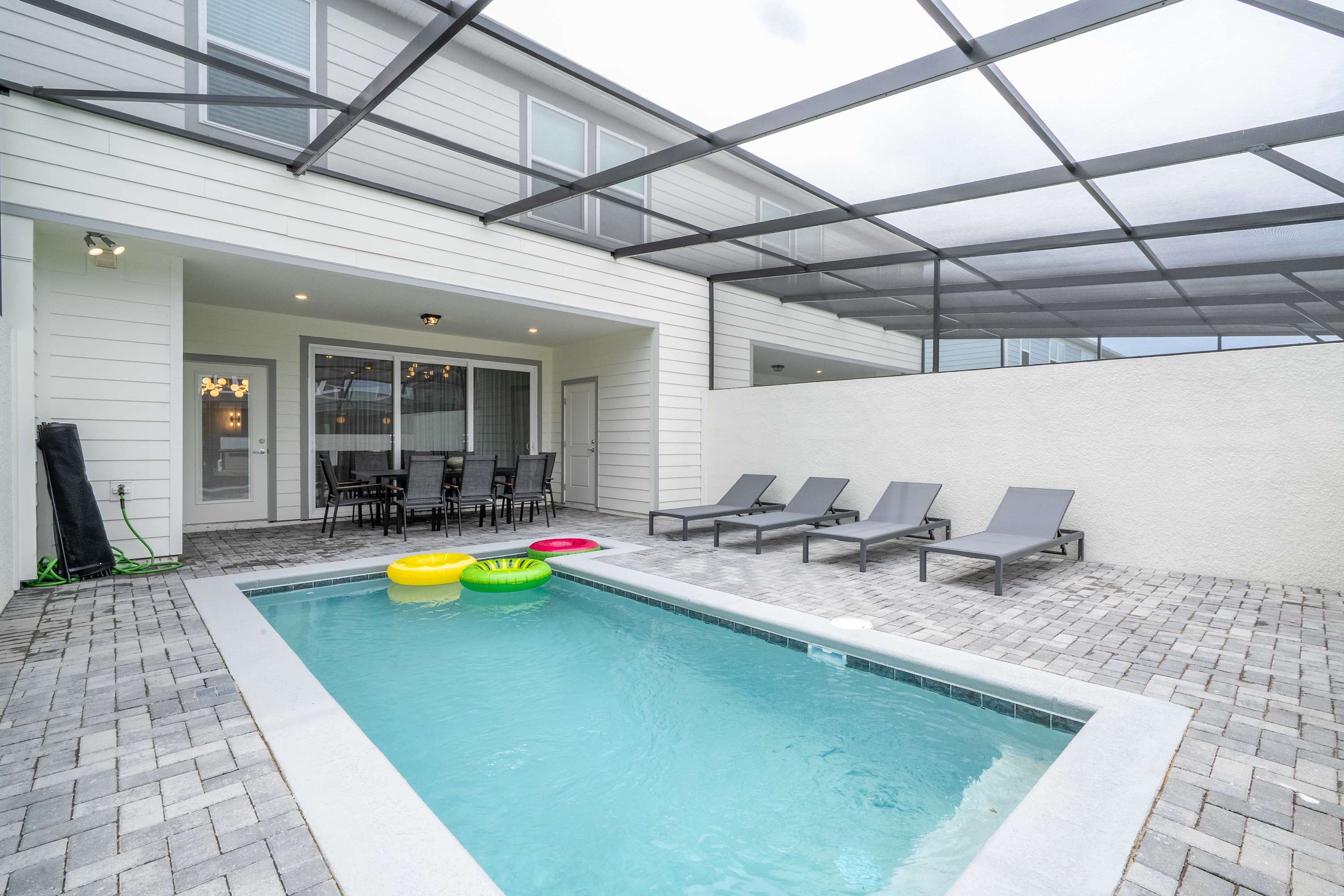 A private swimming pool area, enclosed by a screen cage, features a rectangular pool with colorful inflatable rings and four gray lounge chairs lined against a white wall. The patio is paved with gray bricks, and the house exterior is visible in the background.