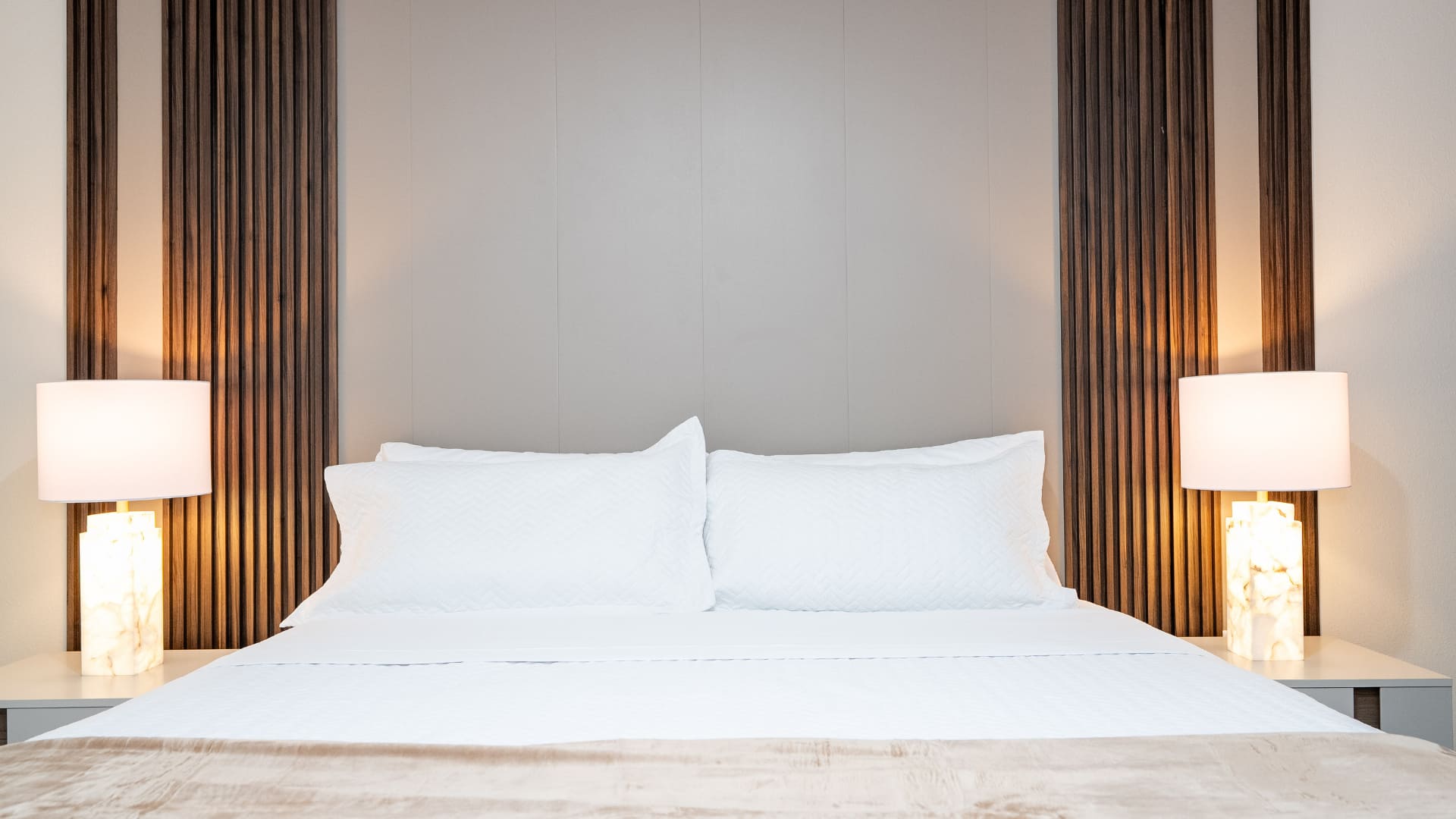A close-up of the bed shows white bedding and pillows with a light brown throw, set against a modern headboard wall featuring vertical wooden panels and a light grey central section. Two illuminated alabaster-style lamps sit on white nightstands.