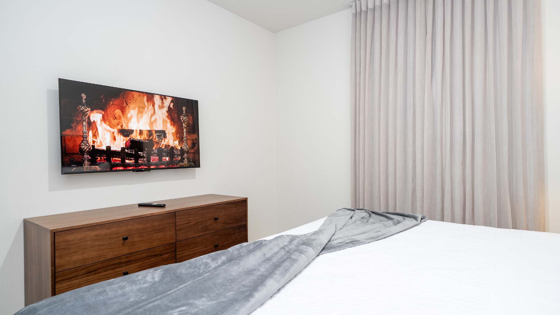 The bedroom features a wall-mounted flat-screen TV displaying a fireplace scene, positioned above a long wooden dresser with multiple drawers. Light grey curtains cover a window on the right, and a grey throw blanket is draped over the bed.