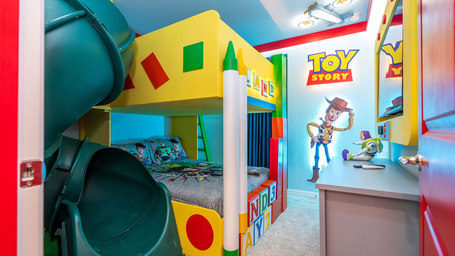 Vibrant Toy Story themed bedroom featuring a yellow and blue bunk bed with a green slide, a large Woody graphic on the wall, and a Buzz Lightyear plush toy.