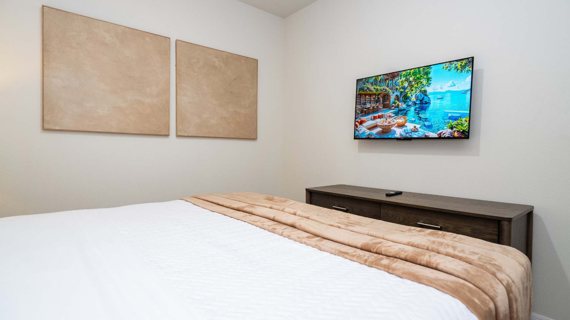 View of a bedroom featuring a wall-mounted flat-screen TV above a dark wood dresser, with a bed covered in white and tan bedding visible in the foreground. Two abstract art pieces adorn the wall.