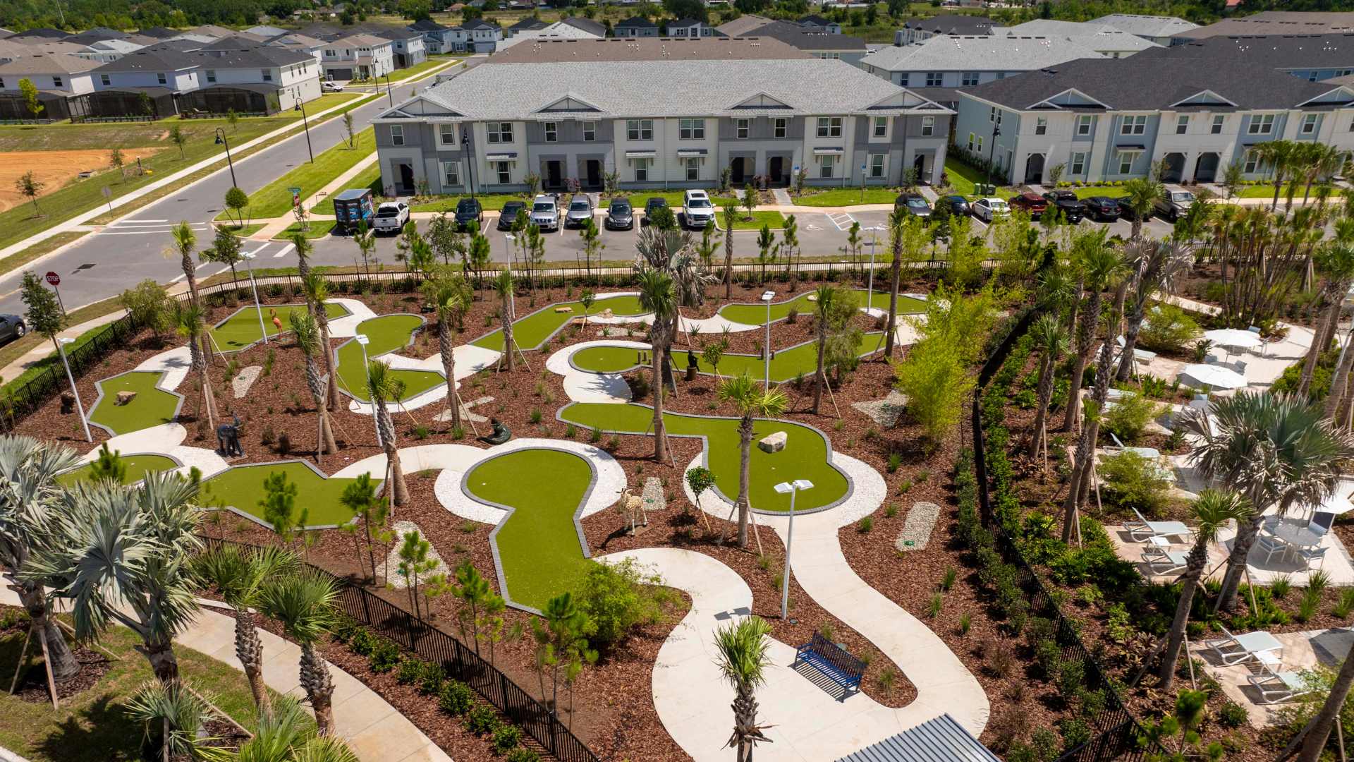 Aerial view of a winding miniature golf course with green turf fairways and white concrete paths, surrounded by mulch, various palm trees, and modern townhouses in the background.