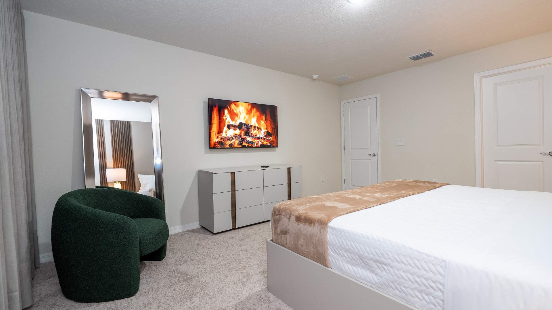 A wide shot of the bedroom shows a wall-mounted TV displaying a fireplace, above a white dresser with wooden accents. A large full-length mirror and a dark green textured accent chair are visible, with light grey curtains covering the window.