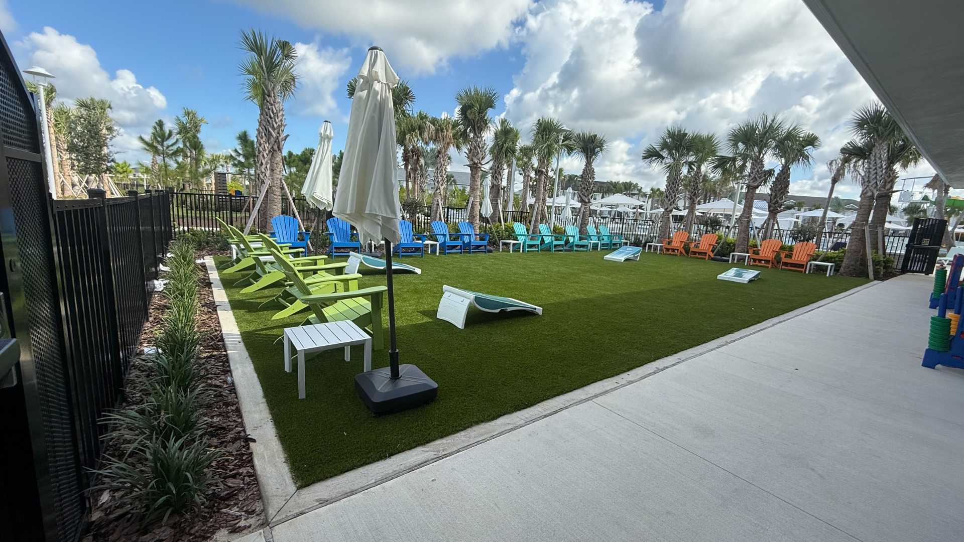 Outdoor recreation lawn with artificial turf, lined with vibrant blue, green, and orange Adirondack chairs, featuring cornhole boards and surrounded by palm trees and a black fence.