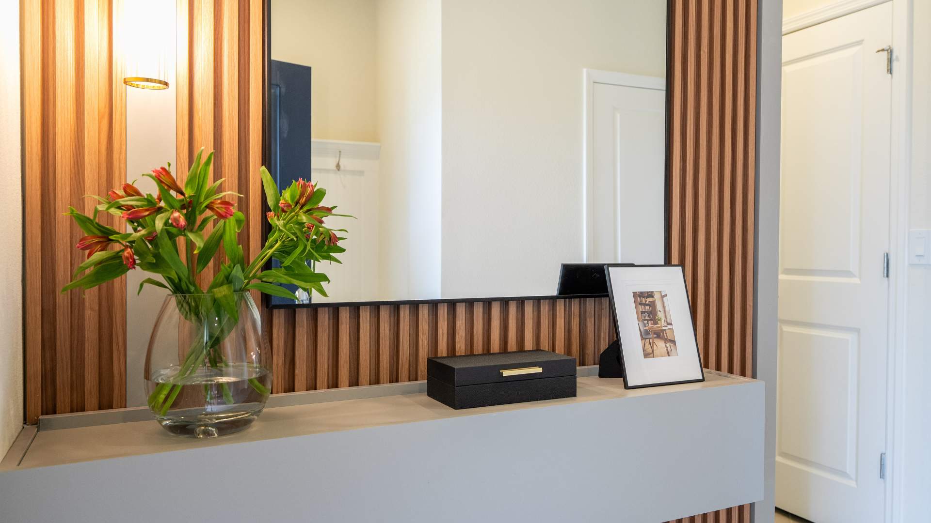 A modern foyer features a light gray console table with a black decorative box and a clear vase of red and green flowers, set against a large mirror framed by vertical wooden slats. A wall sconce with a gold accent provides soft lighting.