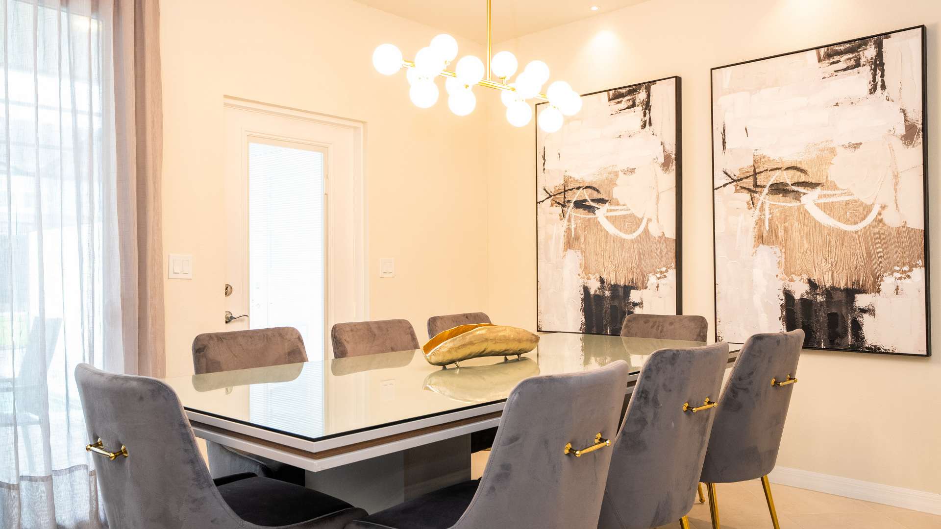 Elegant dining area featuring a rectangular glass-top dining table with a white base, surrounded by eight grey velvet chairs with gold legs. Two large abstract art pieces adorn the wall, and a modern gold chandelier hangs overhead.