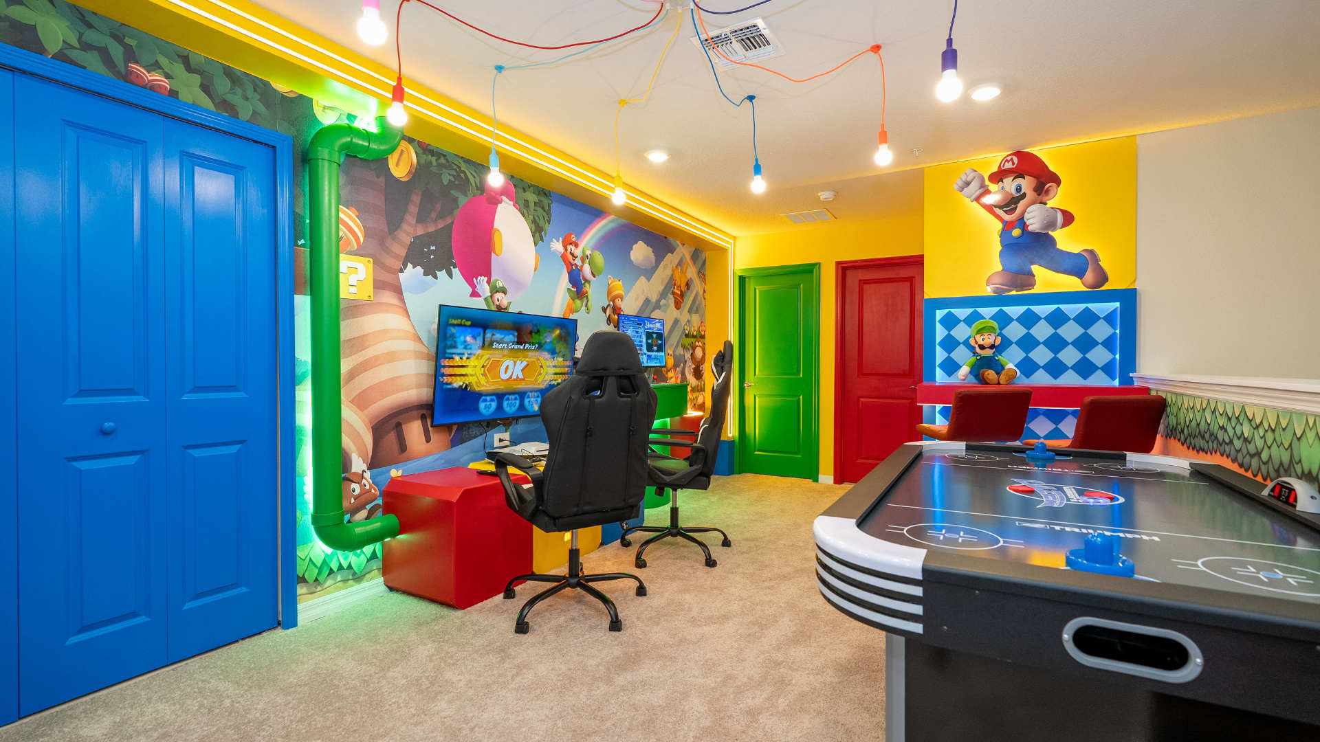 Wide shot of the vibrant Mario-themed game room, featuring two gaming stations with large TVs and black gaming chairs, alongside an air hockey table. The room is adorned with colorful murals, bright yellow walls, and three brightly colored doors (blue, green, red).