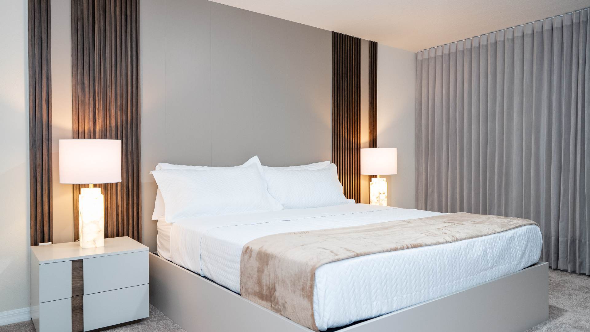A bright bedroom features a large bed with white bedding and a light brown throw, flanked by two white nightstands with illuminated alabaster-style lamps. The headboard wall is accented with vertical wooden panels and a light grey central panel.