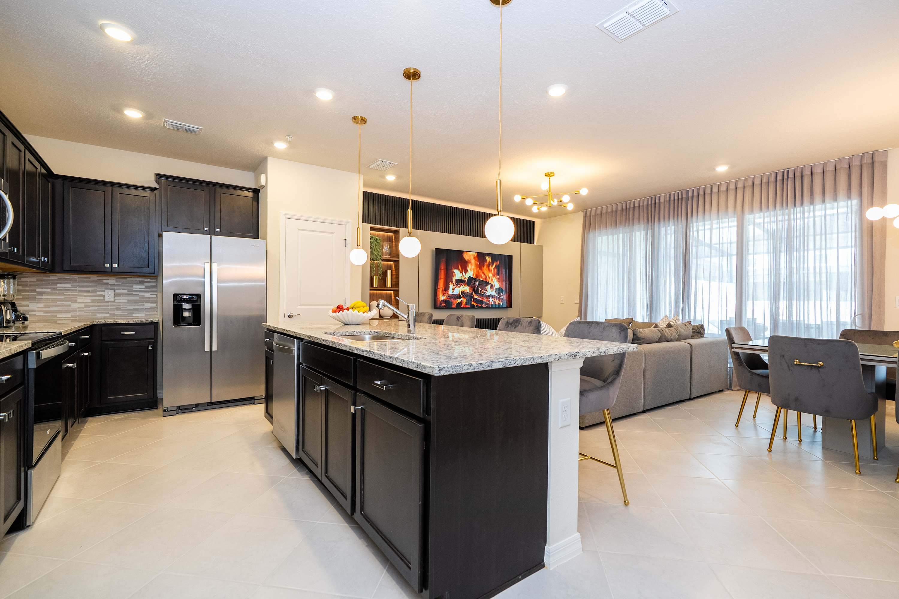 Open-concept kitchen and living area featuring a large granite island with dark cabinets, stainless steel appliances, and gold pendant lights. The living room with a large sectional and a TV displaying a fireplace is visible in the background.