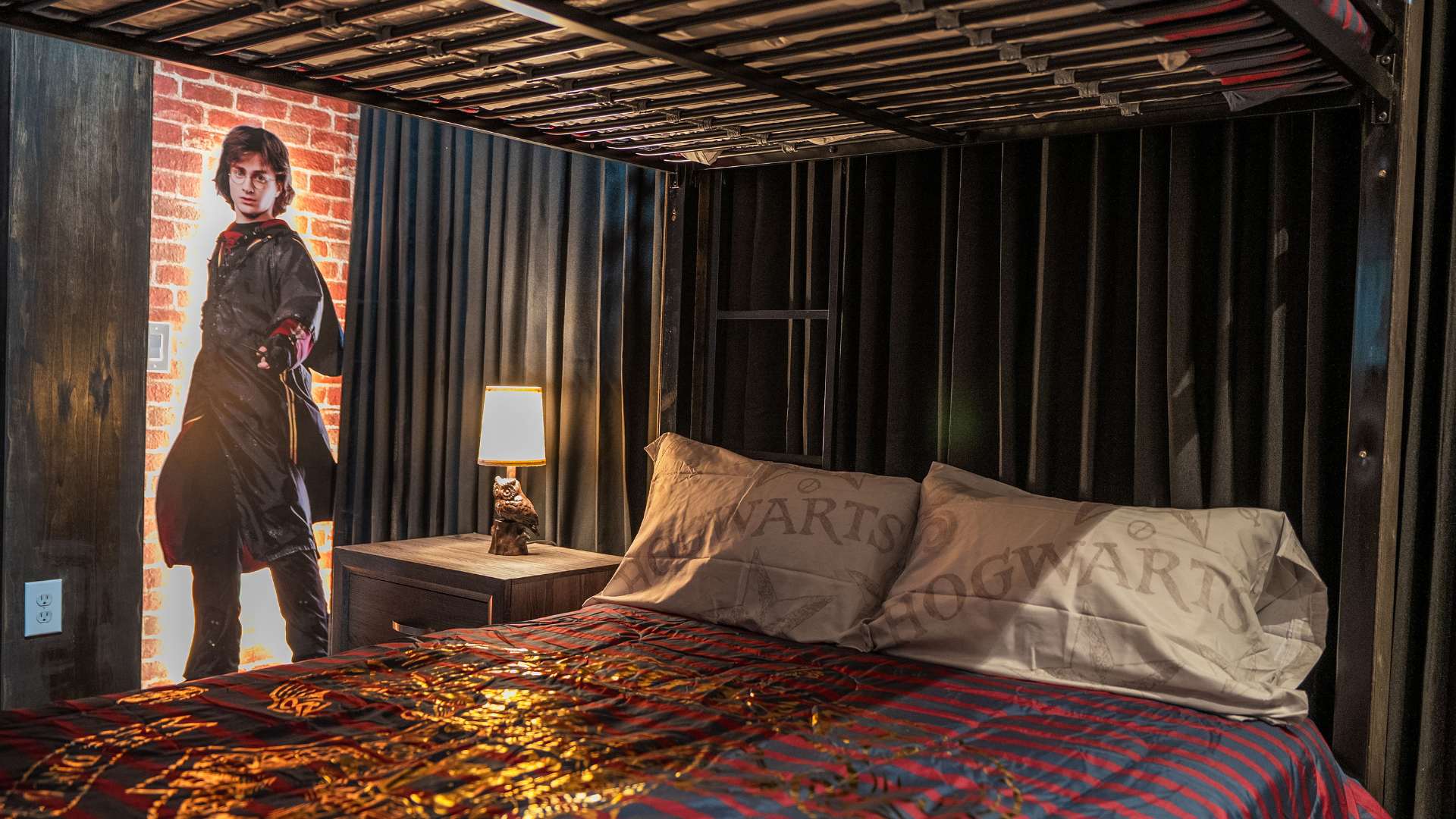 Lower bunk of a Harry Potter themed bedroom with striped bedding, "Hogwarts" pillows, a wooden nightstand with an owl lamp, and a Harry Potter cutout on a brick wall.