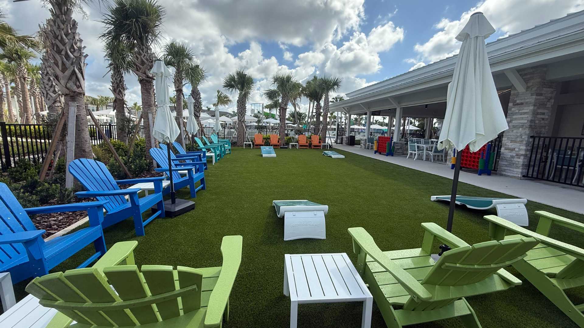 Spacious outdoor recreation area with artificial turf, featuring colorful Adirondack chairs arranged for social gathering, cornhole games, and palm trees, with a covered patio building in the background.