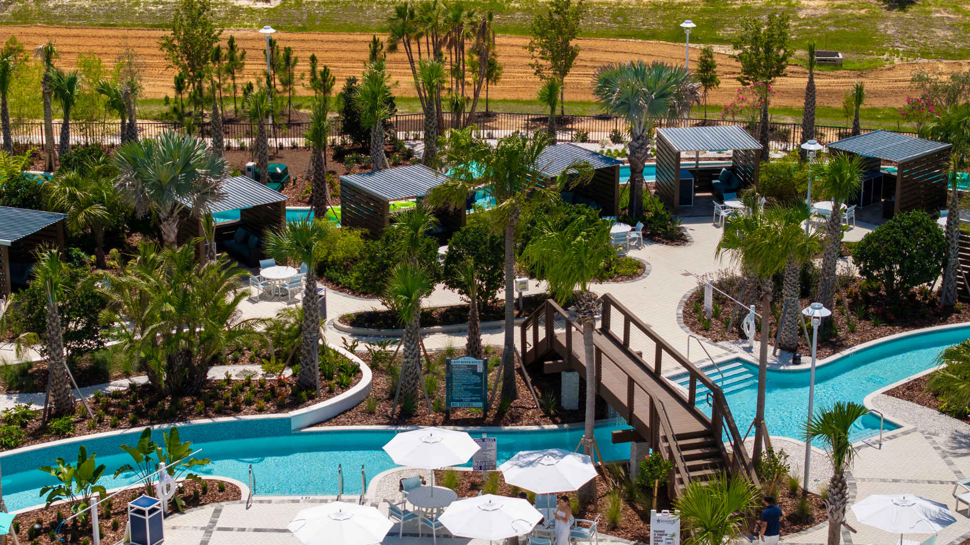 Aerial view of a winding lazy river with clear blue water, surrounded by lush landscaping, wooden bridges, and private cabanas with dark roofs and comfortable seating areas.