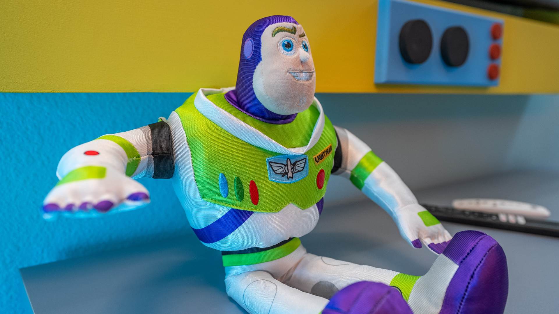 Close-up of a plush Buzz Lightyear toy with a friendly smile, sitting on a blue surface against a yellow and blue background, part of a themed room.