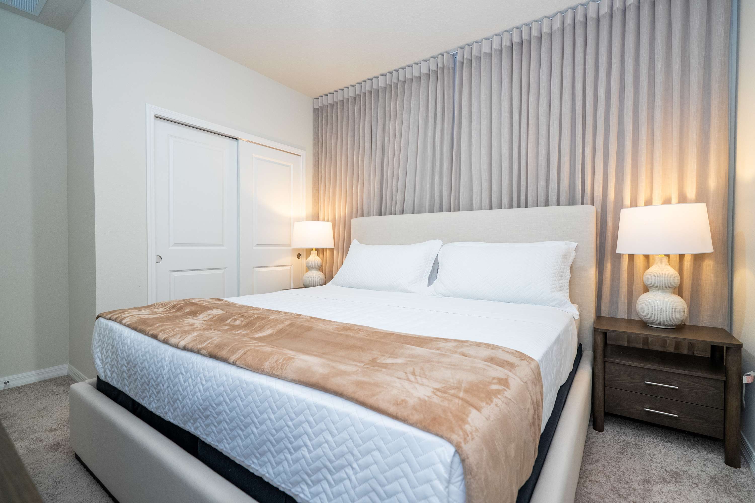 Cozy bedroom with a light upholstered bed, white bedding, and a brown throw blanket. Two nightstands with textured white lamps flank the bed, and light grey pleated curtains cover the wall behind.