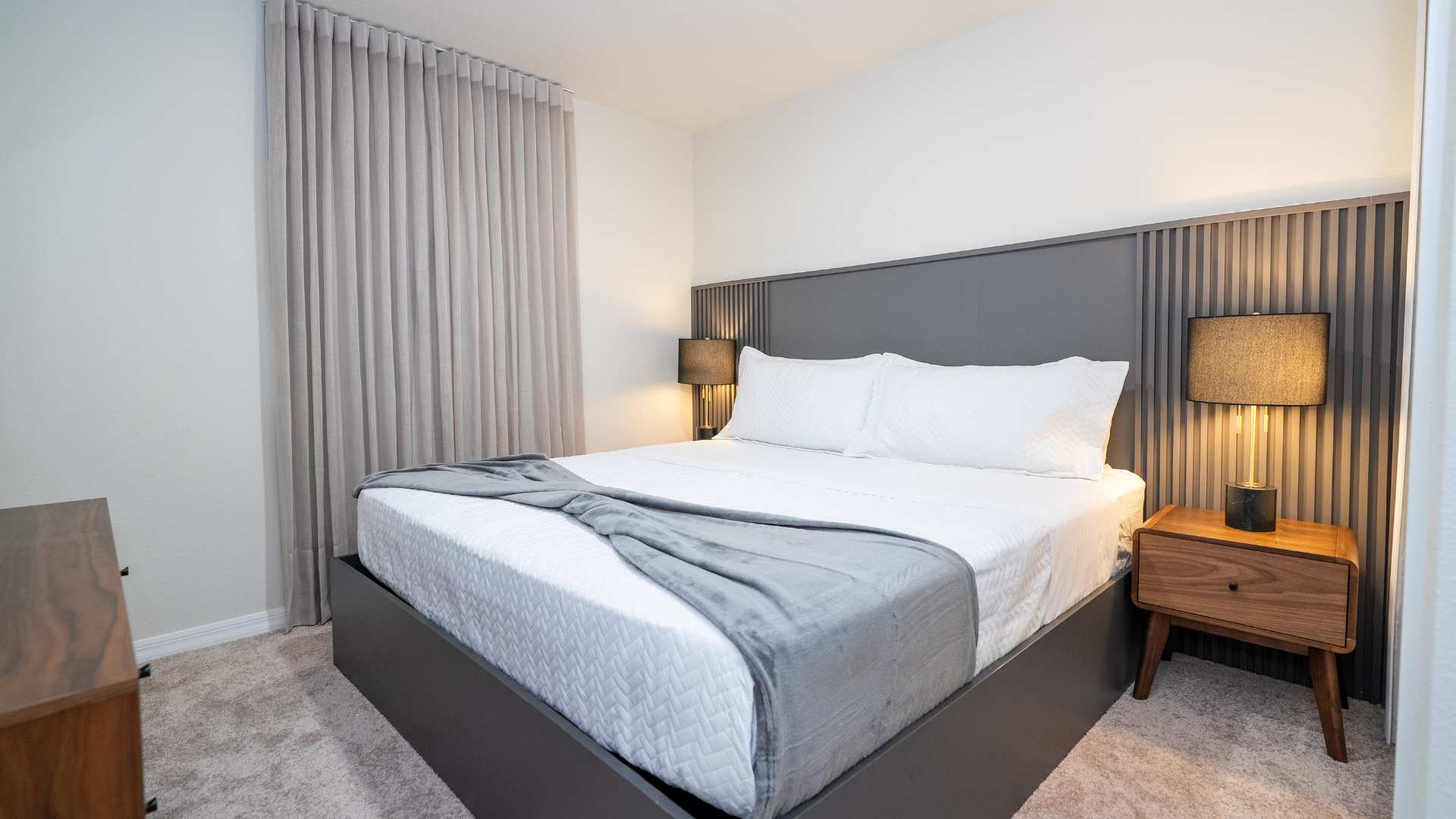 A contemporary bedroom features a large bed with white bedding and a grey throw, flanked by two wooden nightstands with dark lamps. A grey slatted headboard provides a modern accent, and light grey curtains are visible on the left.