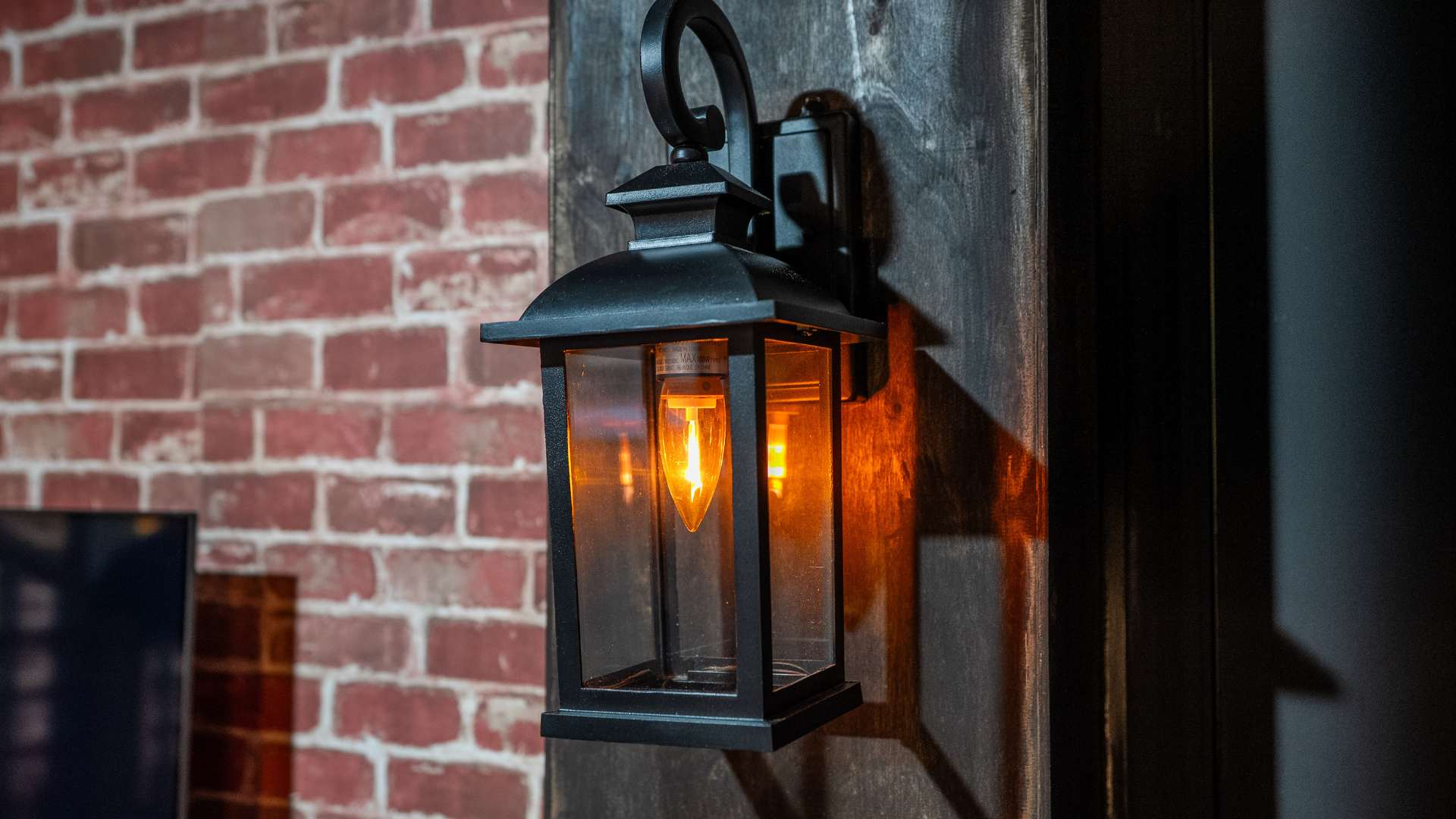 A vintage-style black metal wall sconce with a warm Edison bulb, mounted on a dark wooden beam against a red brick wall, providing ambient light.