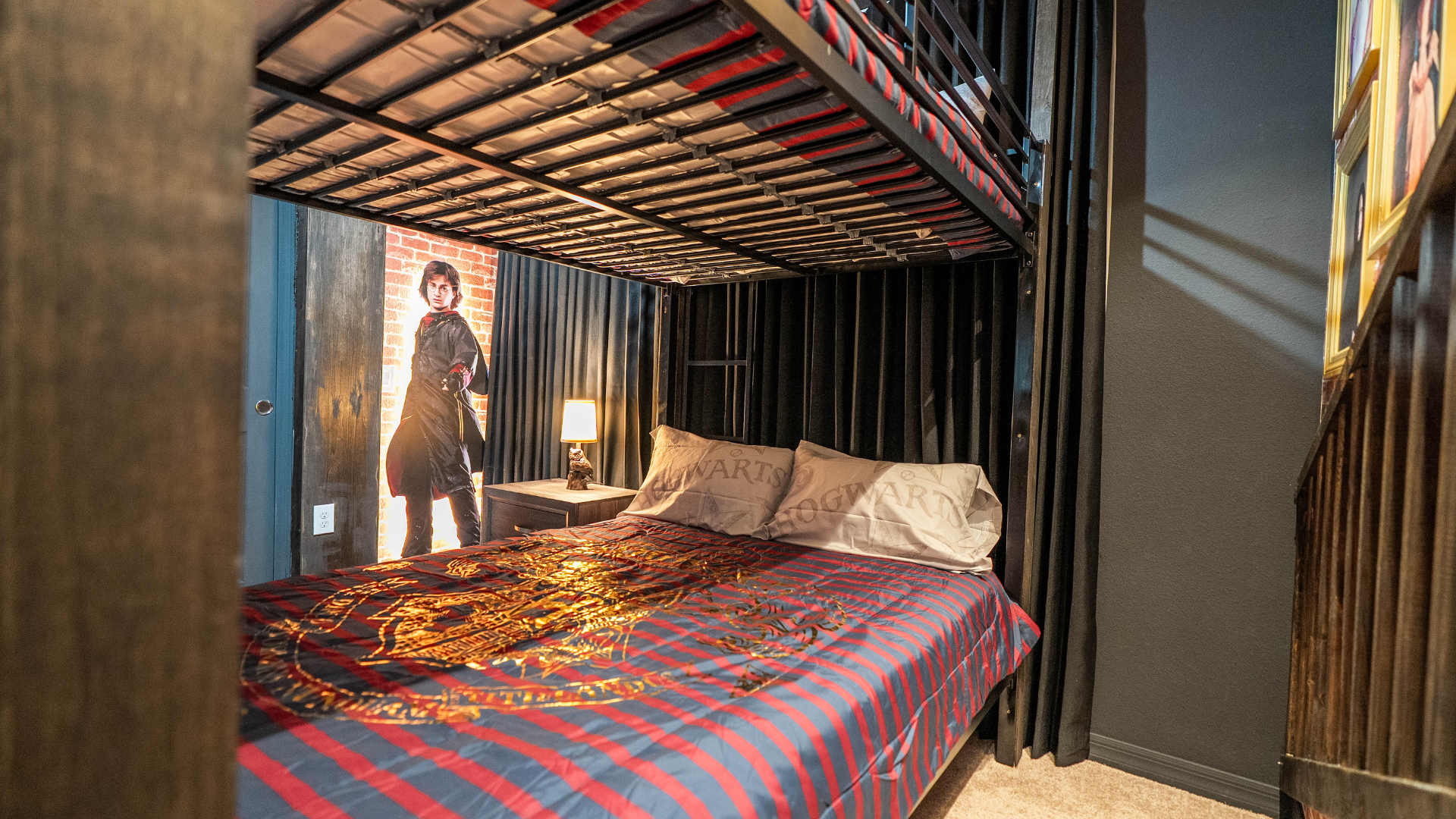 View of the Harry Potter themed bedroom's lower bunk with striped bedding, "Hogwarts" pillows, an owl lamp on a nightstand, and a large Harry Potter cutout against a brick wall.