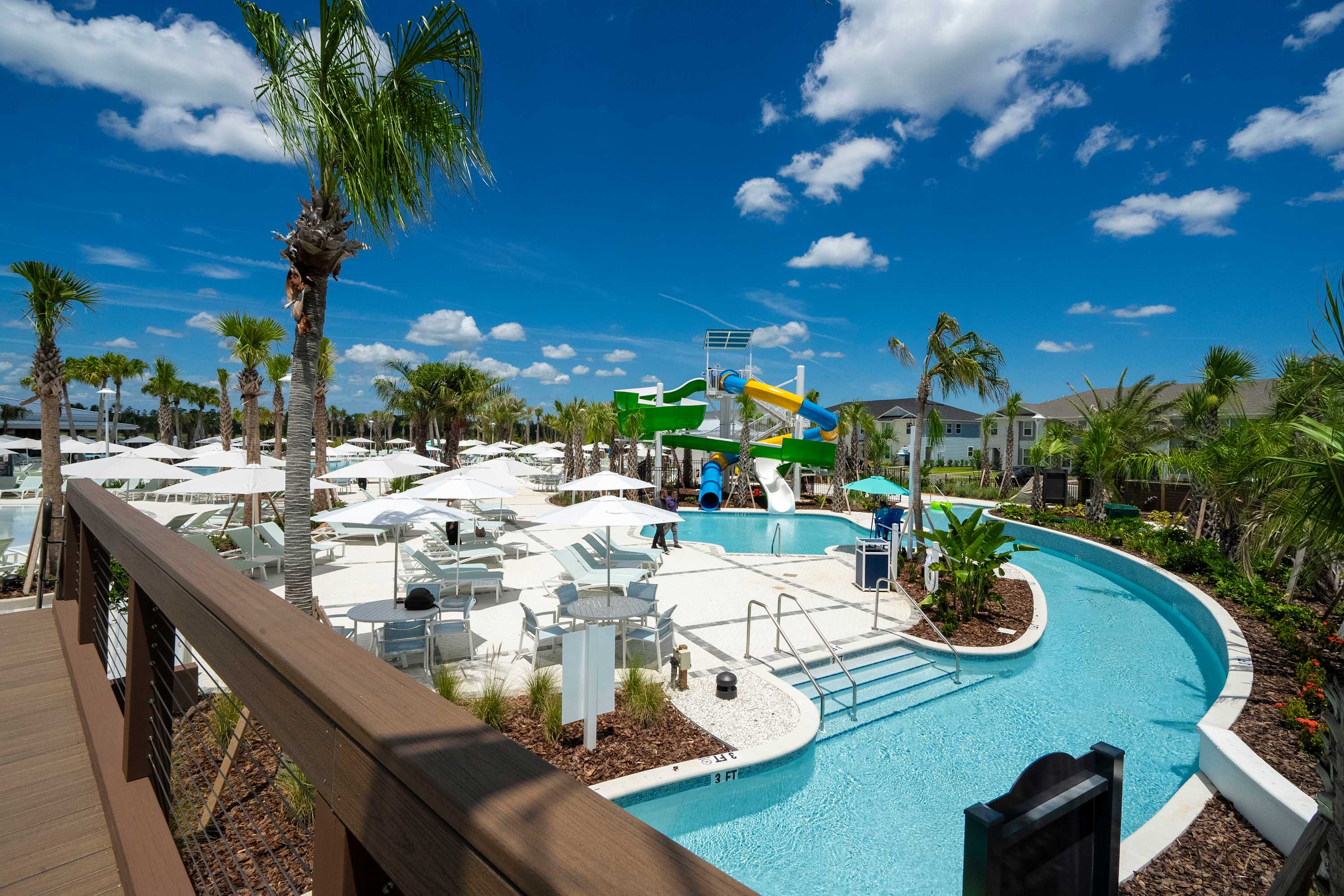 Expansive resort-style pool area featuring a winding lazy river, colorful water slides, and a large deck with numerous white lounge chairs and umbrellas under a bright blue sky.
