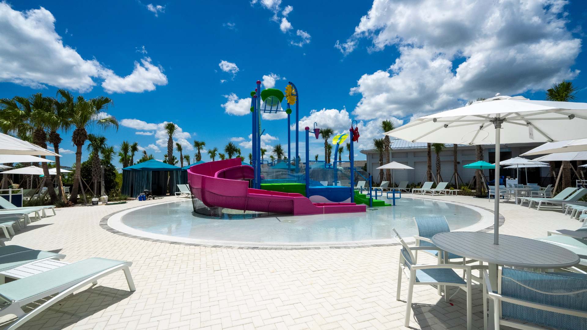 A circular splash pad area designed for children, featuring a vibrant pink water slide and a multi-colored water play structure with buckets and sprayers, surrounded by white lounge chairs and umbrellas.