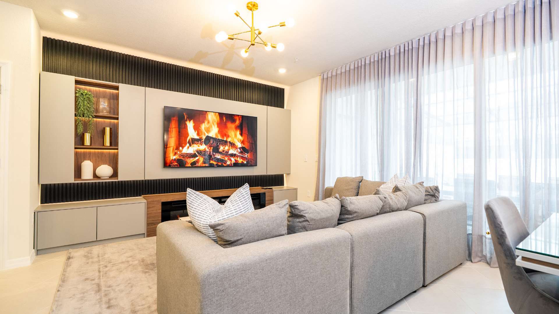 Modern living room with a large grey sectional sofa and a sleek entertainment unit featuring a wall-mounted TV displaying a fireplace scene. The unit has dark slatted accents, built-in shelving with decor, and a long electric fireplace below.