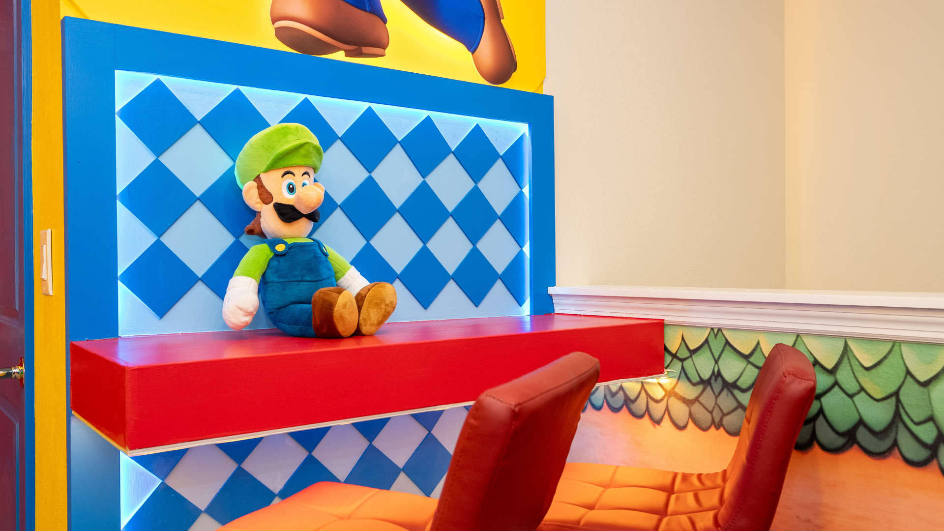 A cozy corner of the game room features a plush Luigi character doll sitting on a red and blue checkered bench with integrated blue LED lighting. Two red gaming chairs are visible in the foreground, complementing the vibrant Mario-themed decor.