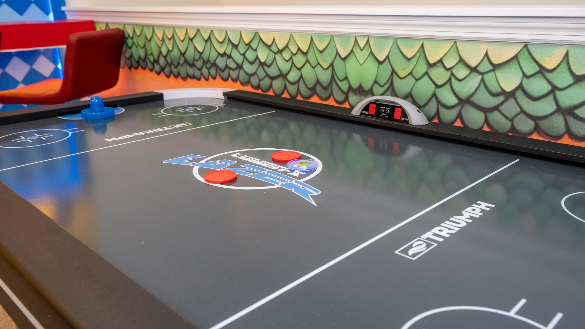 Detailed view of a black air hockey table with red and blue pucks and paddles, featuring the "Triumph" brand logo. The table is positioned against a wall with a green, scaly Bowser-themed mural, enhancing the game room's playful ambiance.