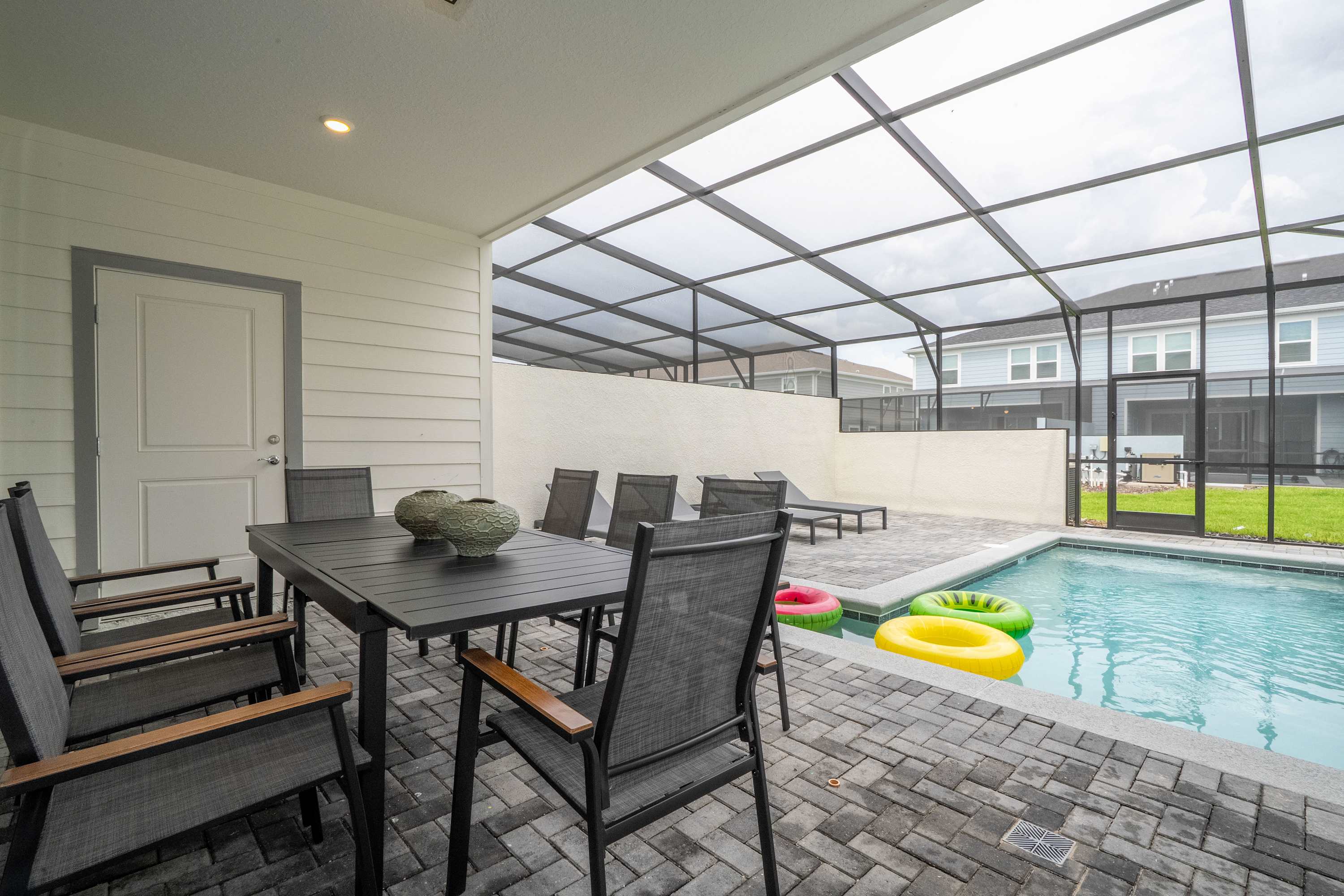 A covered outdoor patio features a dark gray rectangular dining table with six matching chairs, overlooking a small private swimming pool with colorful inflatable rings. The area is enclosed by a screen cage and paved with gray interlocking bricks.