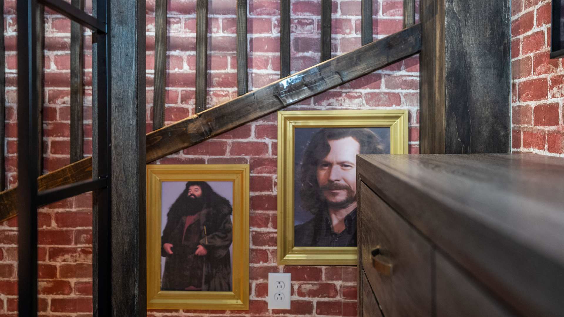 A themed hallway or stairwell area features exposed red brick walls and dark wooden beams, adorned with two framed portraits of characters from Harry Potter, Rubeus Hagrid and Sirius Black. A dark wooden dresser is visible on the right.