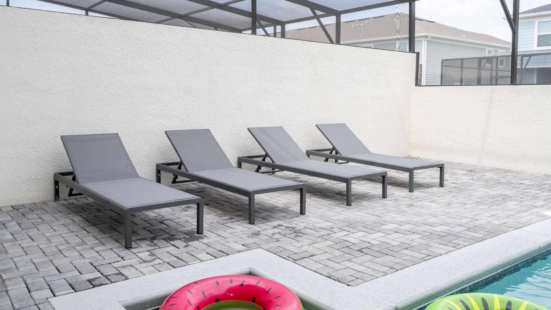 Four sleek gray lounge chairs are neatly arranged on a gray brick patio beside a private swimming pool, with colorful inflatable rings visible in the water. A white textured wall provides a clean backdrop, and a screen enclosure is visible overhead.